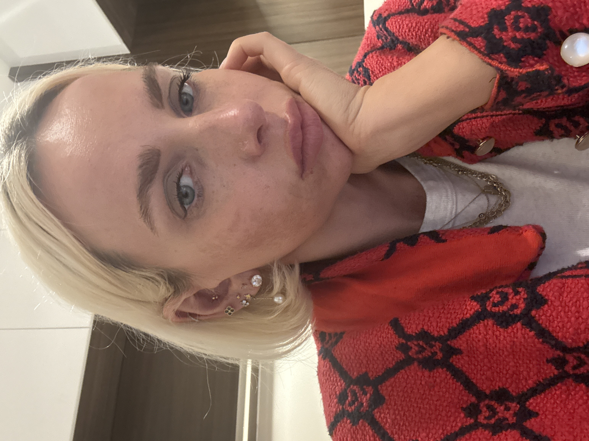 A close-up, indoor selfie shot from above shows a blonde-haired woman with blue eyes. Her left hand is supporting the left side of her face. She is wearing multiple earrings in her left ear. She wears a white t-shirt and a red patterned jacket with large pearl-like buttons. A layered gold chain necklace is also visible. The background is blurred and indistinct, showing what appears to be white wall and a brown-tone cabinet. The photo does not allow to conclude anything about the city of Dubai.