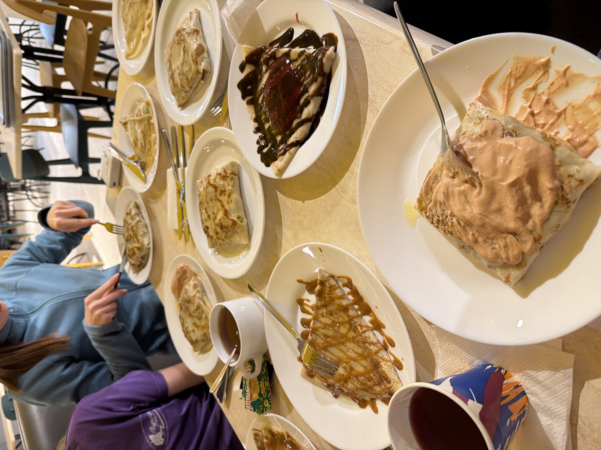 A high-angle shot shows a table laden with plates of crepes and drinks. A person is visible to the left, wearing a blue jacket and purple shirt, holding cutlery. Ten plates containing folded crepes with various toppings (including chocolate, caramel, and a pinkish-brown sauce) are arranged across the table. Several cups containing liquid are also present, and cutlery is placed beside each plate. The scene appears to be indoors, possibly in a restaurant. There is no definitive information in the image to confirm its location as Berezniki, Russia.
