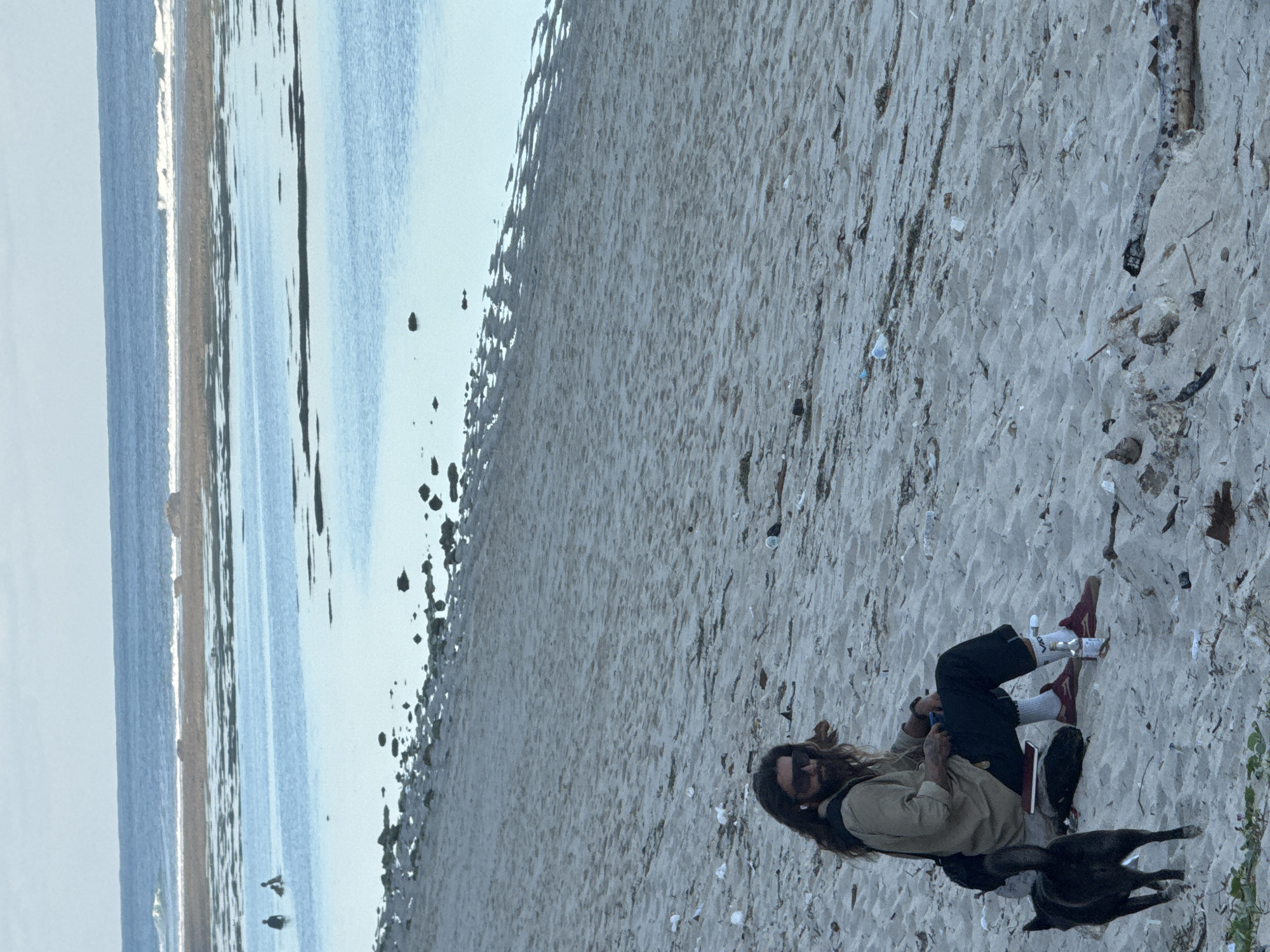 A man with long brown hair and sunglasses sits on a sandy beach in Kuta Selatan, Indonesia. He is wearing a tan shirt, black pants, white socks, and burgundy sneakers. A black dog stands to his left. In the background, the ocean is visible with a shallow, blue water area that recedes to a darker, rougher sea with waves. Several rocks are visible in the water. The beach itself is covered in sand with scattered debris, including plastic bottles and driftwood. Two figures are visible in the water in the distance, appearing to be wading.