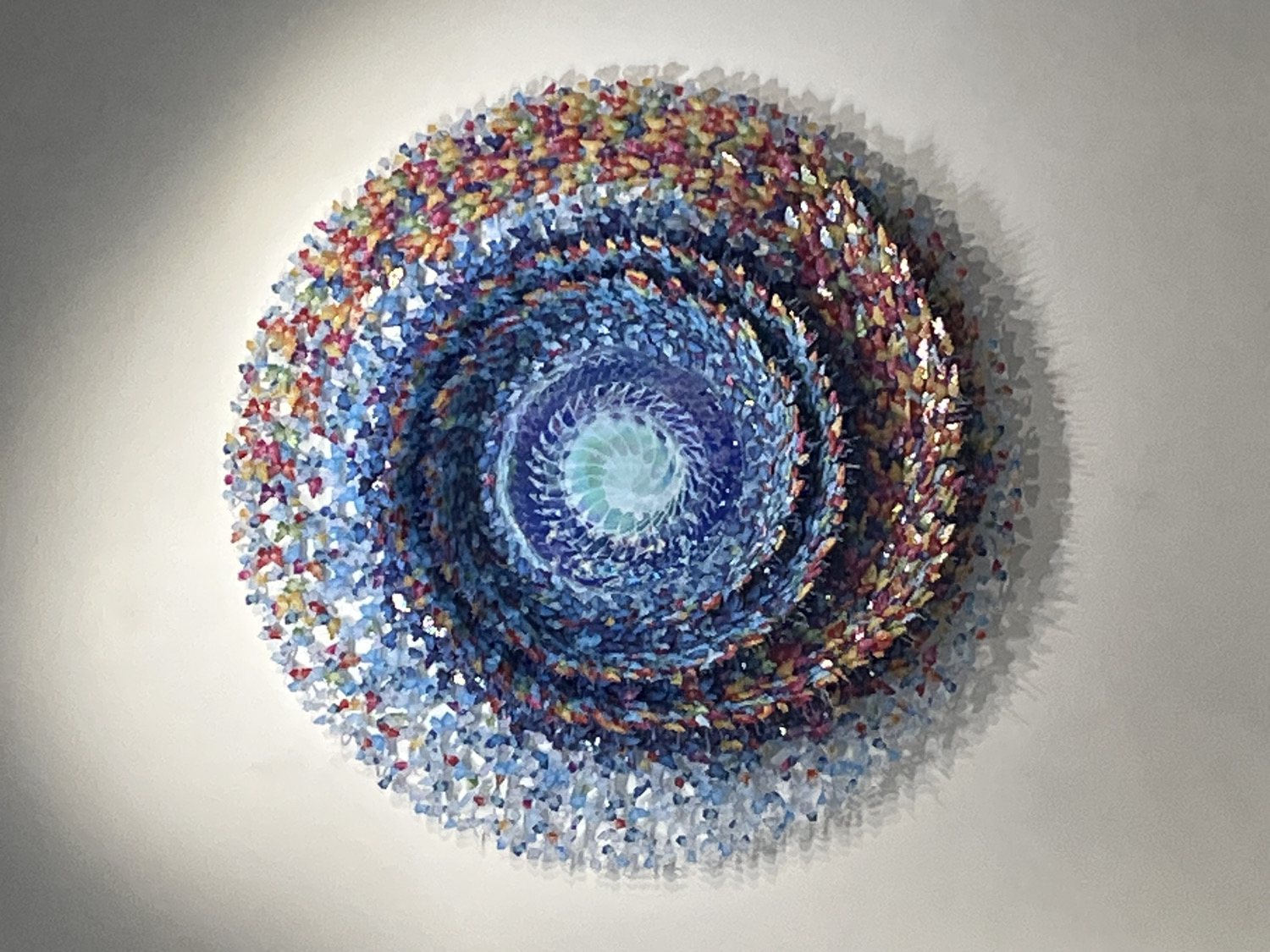 A vibrant, three-dimensional circular artwork is prominently displayed against a plain, light-colored wall, in what is contextually known to be Barcelona, Spain. The artwork is meticulously crafted from countless small, individual pieces, possibly resembling tessellated fragments or delicate petals. These pieces are arranged in a captivating spiraling pattern that emanates from a luminous, pale blue and turquoise core. As the spiral expands outwards, the colors dramatically transition through a spectrum of deeper blues, rich purples, and vibrant reds, culminating in warm oranges and bright yellows at its outermost edges.
The intricate construction gives the artwork significant depth, with the numerous small components appearing to be slightly raised from the wall. This three-dimensional quality is accentuated by the shadows cast upon the wall behind the piece, highlighting its relief and textural complexity. The lighting, likely artificial, brightly illuminates the artwork, making its diverse colors and detailed composition stand out. There are no people or specific activities depicted in the image. The scene is a static presentation of this elaborate, abstract artwork. No visible text is present within the frame.