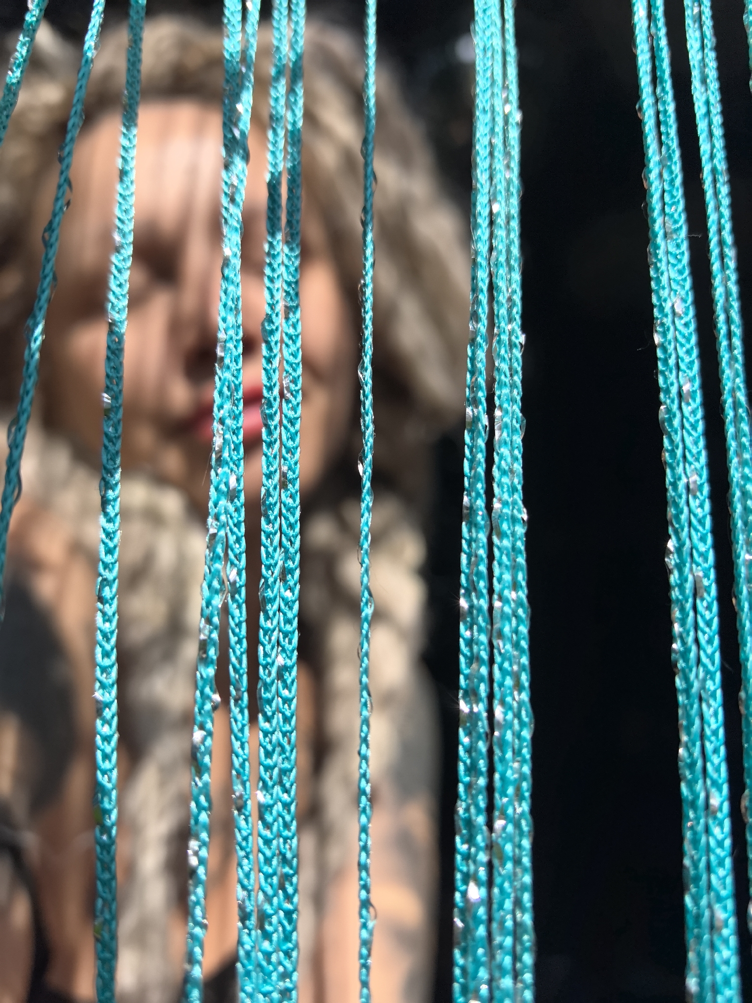 The image features a close-up view of several vertical strands of braided teal or turquoise cord, sharply in focus and positioned in the foreground. These cords are adorned with numerous small, clear, reflective droplets, likely water or decorative beads, which glisten under bright light.
Behind the prominent cords, the blurred figure of a person is visible in the mid-ground. The person appears to be a woman, with light-colored, possibly tousled or curly hair, and distinct red lips, though her eyes are obscured by the blur and the foreground cords. She seems to be wearing a textured, possibly furry, collar or garment around her neck and shoulders. On the left side of the frame, a blurred arm with what appears to be a tattooed pattern is also discernible.
The background behind the person is predominantly dark, almost black, creating a strong contrast with the brightly lit foreground elements. The bright reflections on the droplets suggest illumination from a strong light source, possibly sunlight. The overall scene gives the impression of observing the person through a decorative curtain or screen made of the beaded cords. No specific activity is clear, and the emotions of the subject cannot be determined due to the extreme blur and partial obstruction of her face. There is no visible text in the image.