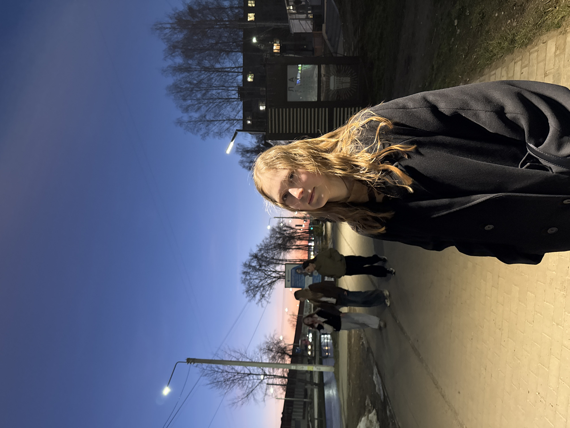 A woman with blonde hair, wearing a black coat, stands on a sidewalk in what appears to be the city of Chaikovsky, Russia. In the background, three other people are walking on the sidewalk. Streetlights illuminate the scene, and a building is visible in the background on the left. The sky transitions from blue to orange-pink towards the horizon.