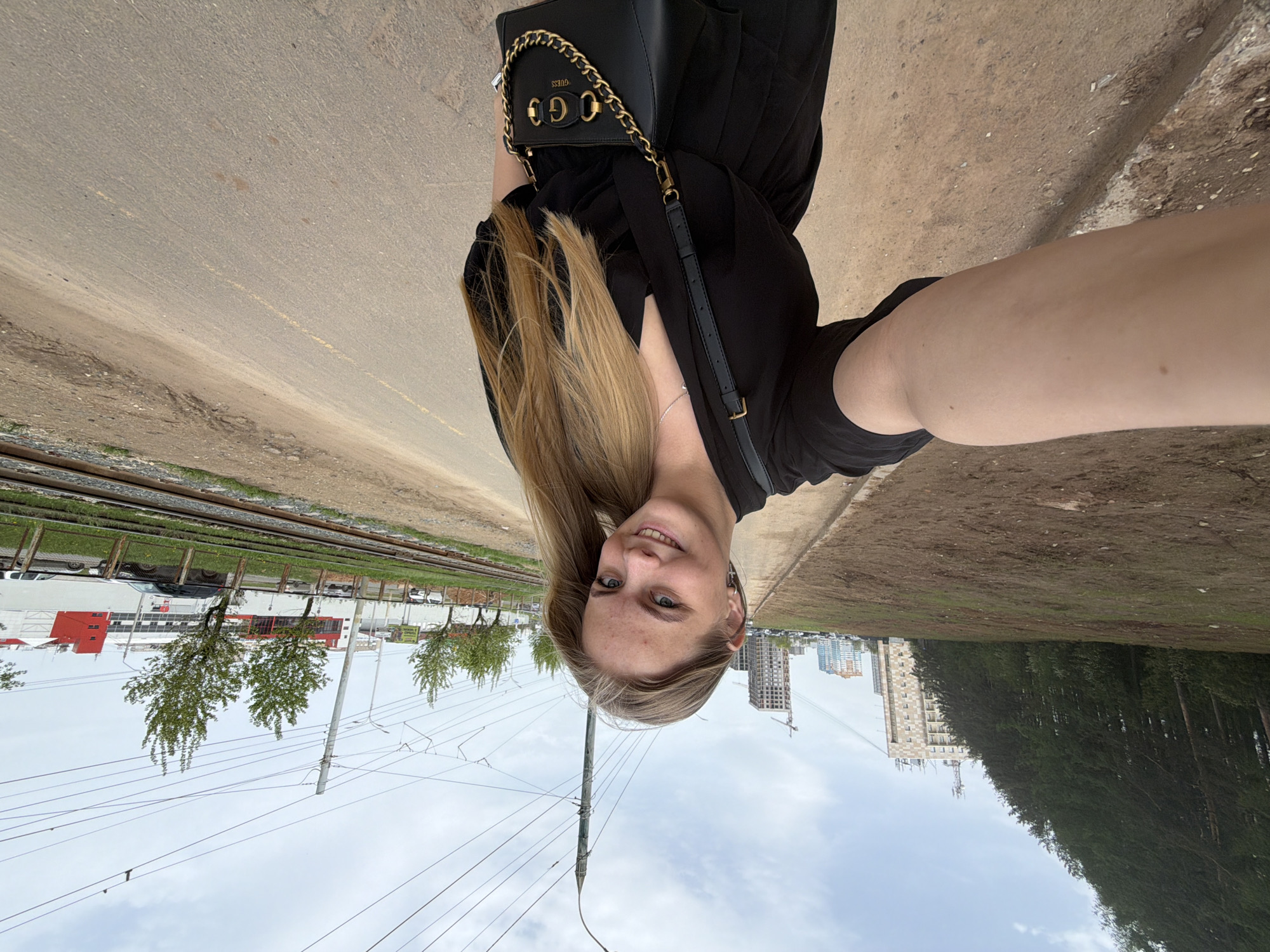 A woman is posing for a selfie while holding the camera at an angle. She has long, blonde hair and is wearing a black dress with a black handbag over her shoulder. The photo appears to be taken outdoors on a path adjacent to a railway track and a grassy area. In the background, there is a cityscape with several tall buildings, including one under construction with a crane. Based on the architecture and urban layout, this image was taken in Izhevsk, Russia. A forest lines the right of the image, and the sky is overcast. Electrical wires and a utility pole are prominently featured between the woman and the background cityscape.