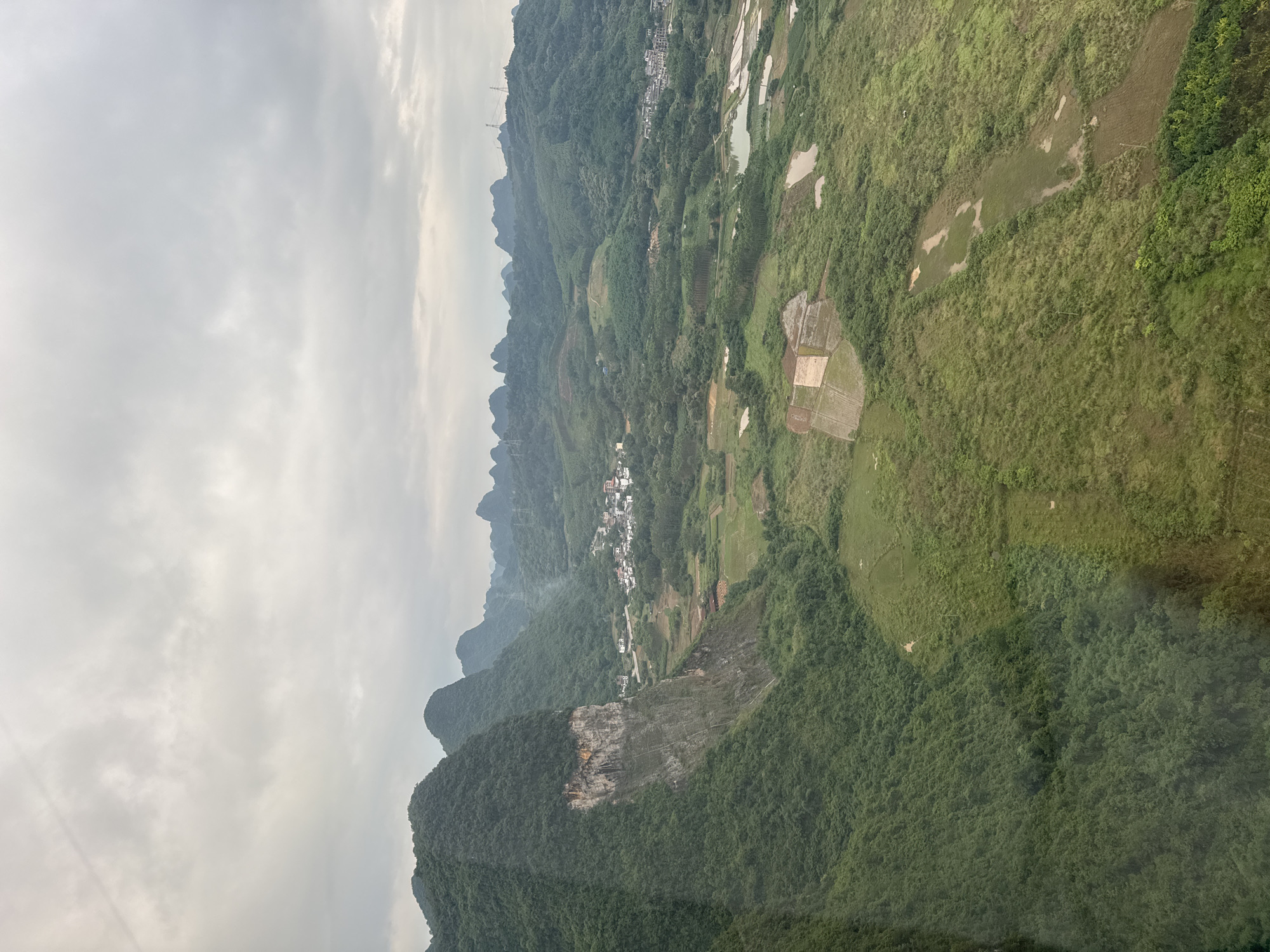 The image shows an aerial view of a landscape near Guilin, China. The scene features karst mountains with dense green vegetation. A portion of one mountain appears to be a gray, exposed rock face. In the valley, there is a small village nestled amongst the hills. Agricultural land with paddies and fields are visible. The sky is overcast with gray clouds.