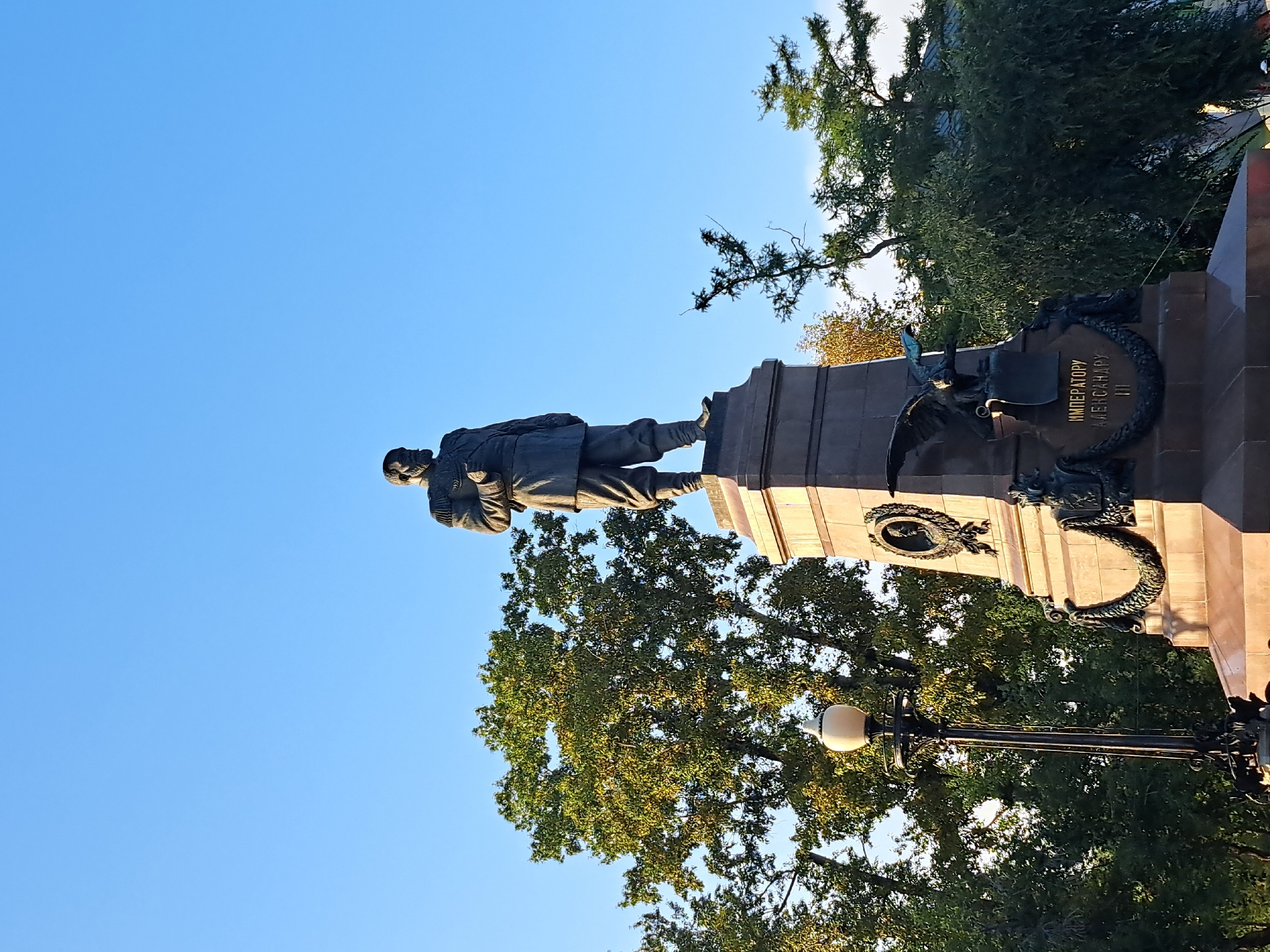 The image shows a bronze statue of a man in military uniform standing on a stone pedestal. The statue is located in Irkutsk, Russia, specifically on Kirov Square. The man in the statue is Emperor Alexander III, dressed in a uniform with epaulets and a sash. He is depicted with a beard and mustache, and his right hand is resting on his chest.
The pedestal is adorned with decorative elements, including a wreath and an eagle sculpture. Inscriptions in Russian are visible on the pedestal, reading "ИМПЕРАТОРУ АЛЕКСАНДРУ III" (To Emperor Alexander III). A lamppost with an ornate black metal base and a white glass lantern is positioned to the left of the statue.
The background consists of a clear blue sky and lush green trees, suggesting an outdoor setting, likely a park or public square. The lighting indicates it is daytime, possibly late afternoon or early morning, given the quality of light. The overall scene is a static portrayal of a historical monument within an urban environment.