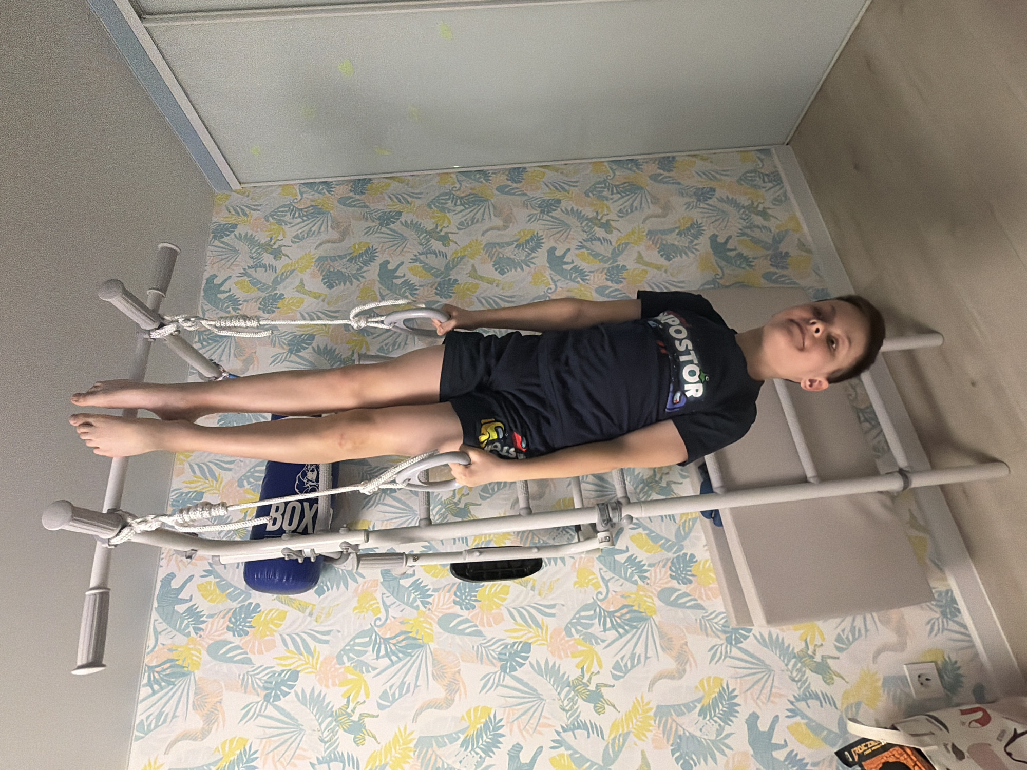 A young boy is lying on a white and gray Swedish ladder or wall bars, commonly found in homes and gyms for exercise. The ladder is positioned against a wall covered in a light blue and yellow patterned wallpaper featuring what appears to be leaves and animal figures. He is wearing a dark-colored t-shirt and shorts. The boy is holding onto suspension rings attached to the ladder. A blue punching bag with white lettering is visible near the ladder. The image was taken in Votkinsk, Russia.