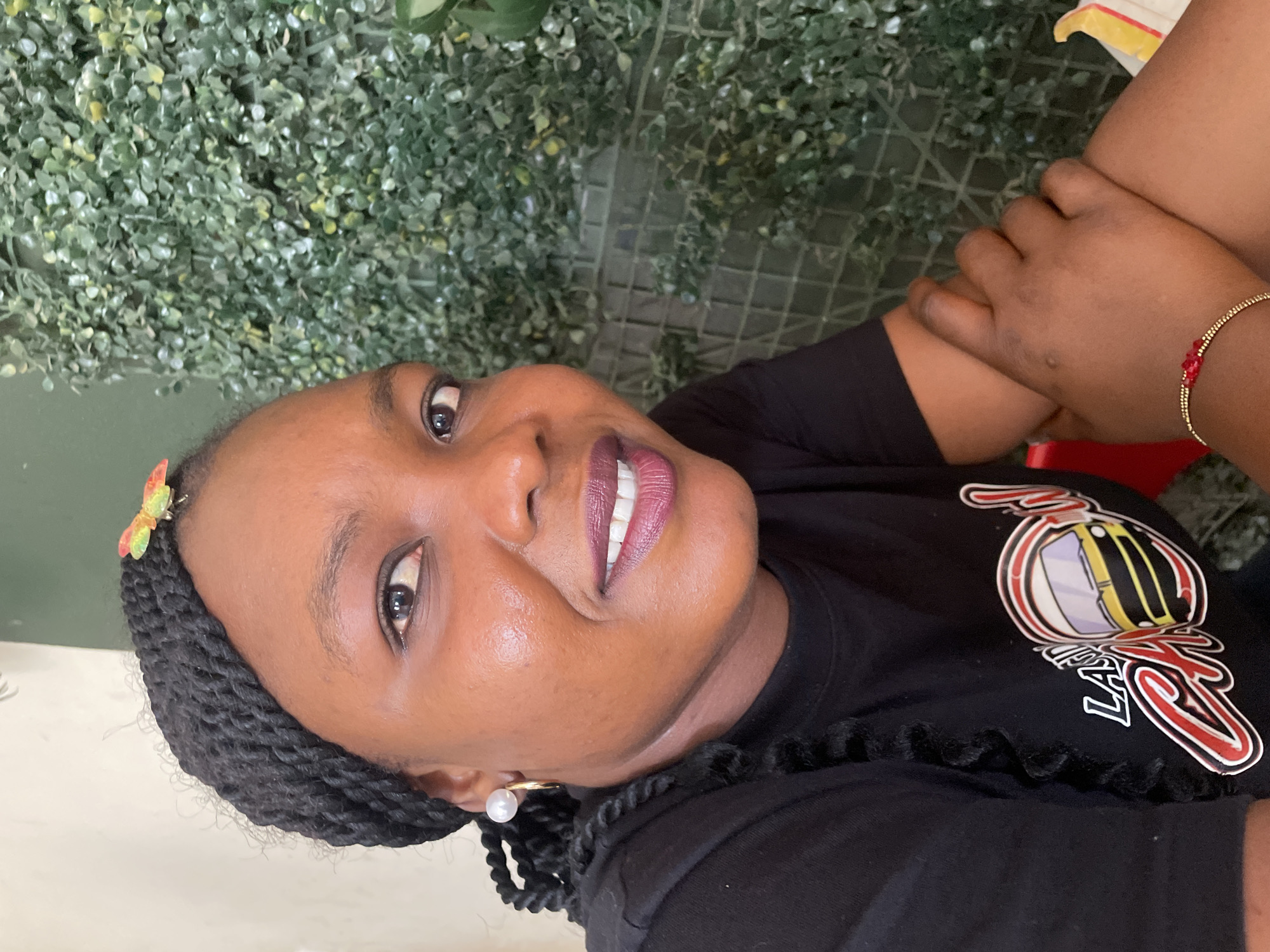 A woman with dark skin and braided hair is looking upwards with a slight smile. She is wearing a black t-shirt with a graphic print in white, red, and yellow. A pearl earring is visible on her left ear and a colorful butterfly hair clip is in her hair. She is wearing a gold beaded bracelet with red beads on her right arm. Her hands are clasped together. The background consists of artificial green foliage. There is no specific indicator of Jos, Nigeria in the image.