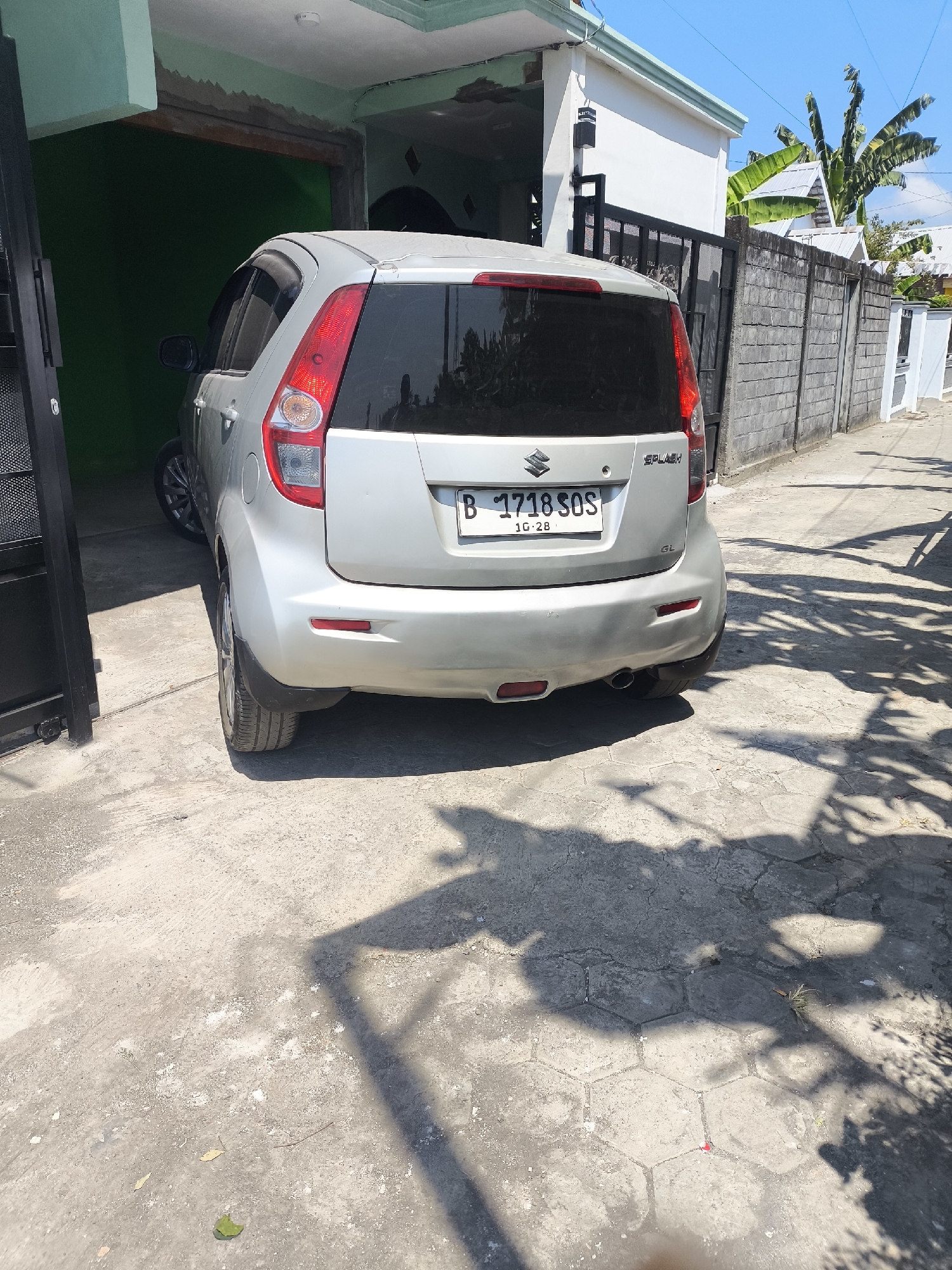 A silver Suzuki Splash car is parked in front of a house on a sunny day in Lumajang, Indonesia. The car is viewed from the rear and is partially covered by the shadow of a tree. The license plate reads "B 1718 SOS". The house has a green painted wall visible inside a garage opening, and a white exterior wall. A concrete fence runs along the side of the property, with a black metal gate in the foreground. Palm trees and other vegetation are visible in the background, along with a clear blue sky. The ground is paved with concrete tiles.
