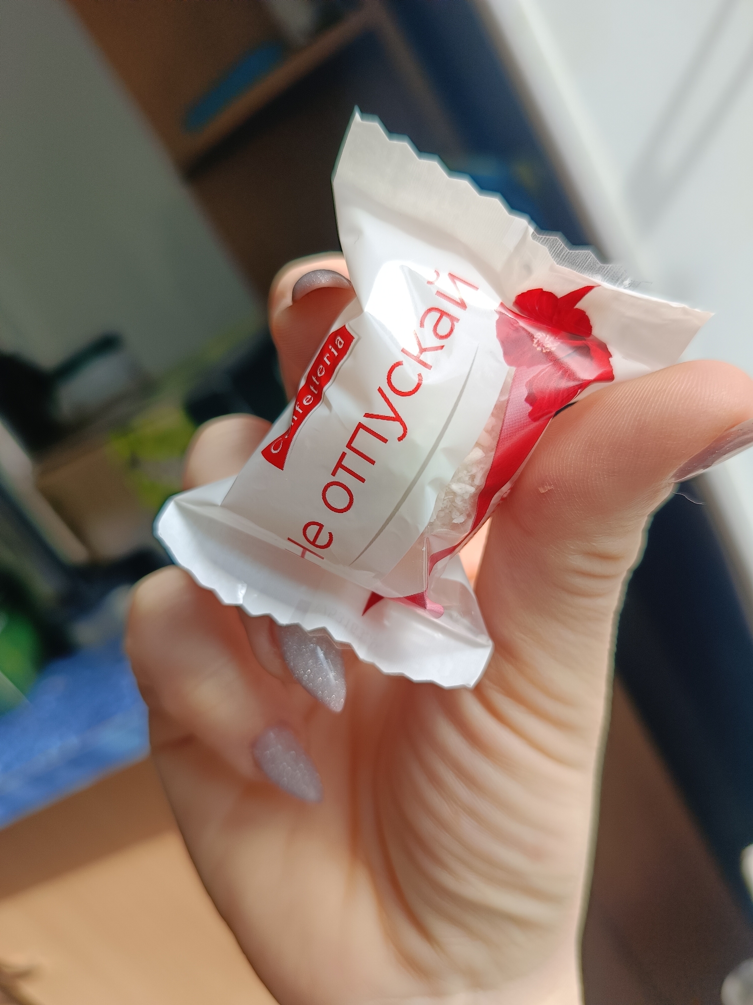 A hand holds a wrapped candy. The candy's wrapper is white with red text in Russian. The word "Cafetteria" is visible. The nails on the hand are painted with a light grey, sparkly polish. The background shows a blurred room with a shelf and other objects.
