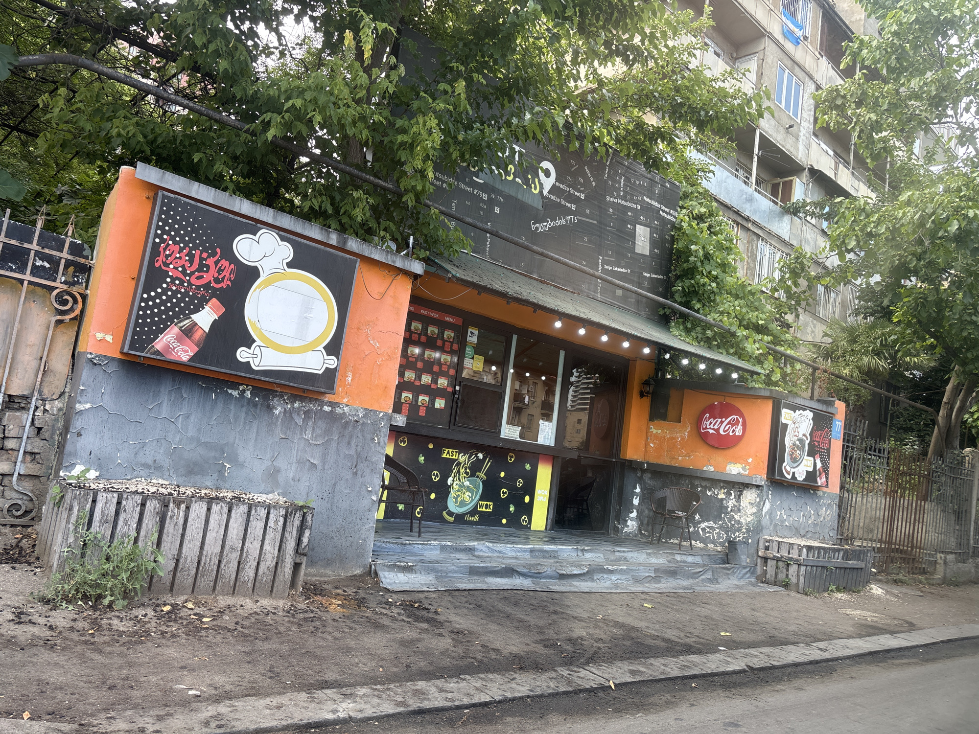 This is a medium-shot, slightly angled outdoor photograph of a street-level food establishment in Tbilisi, Georgia. The building's facade is a mix of weathered concrete and vibrant orange paint, with a large black sign featuring text in Georgian and English, as well as graphics of a Coca-Cola bottle and a chef's hat. Below this sign, the shop's entrance is flanked by windows showcasing menu items, and the word "FAST WOK" is prominently displayed. Further along the facade, a circular Coca-Cola sign is visible. The shop is decorated with a mural of a wok filled with noodles and other ingredients, alongside small pretzel-like graphics. Outdoor seating with chairs is visible on either side of the entrance.
The setting appears to be a somewhat urban environment, with a multi-story apartment building visible in the background, partially obscured by lush green trees. The ground in front of the establishment is paved, with some dirt and debris. A wooden planter box filled with some sort of organic material is situated to the left of the entrance. A metal fence runs along the right side of the image. The time of day appears to be daytime, with bright, natural lighting suggesting clear weather. The overall impression is that of a casual, no-frills eatery. The visible text confirms that the location is Tbilisi, Georgia, with street names like "Nutsubidze Street" and "Tavadze Street" mentioned on the signage.
