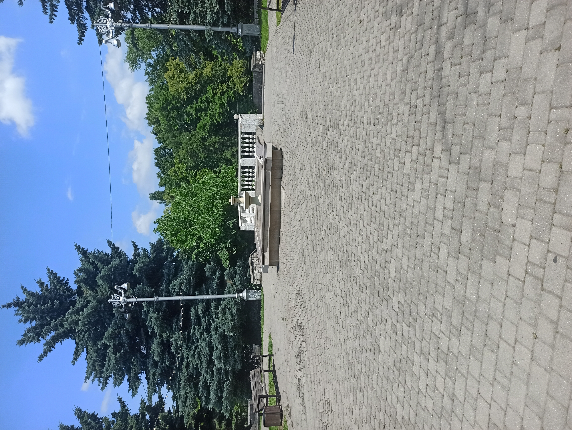 The image depicts a paved area in a park under a bright, cloudy blue sky. In the foreground, a gray, interlocking paver pathway leads towards a central area where a fountain is visible. The fountain appears to be a multi-tiered structure made of stone, with a small spout at the top. It is surrounded by a low, white balustrade with ornate pillars. To the left of the fountain, a wooden bench with a trash can next to it is situated on a patch of grass.
Further back, a line of tall evergreen trees, possibly firs, dominates the background, with dense green foliage. A traditional lamppost with a decorative metalwork top and a single light fixture stands near the fountain. Another similar lamppost is visible in the far right of the image, partially obscured by trees. Beyond the evergreen trees, a variety of deciduous trees with lush green leaves form a dense canopy. The overall scene suggests a well-maintained public park or garden area.