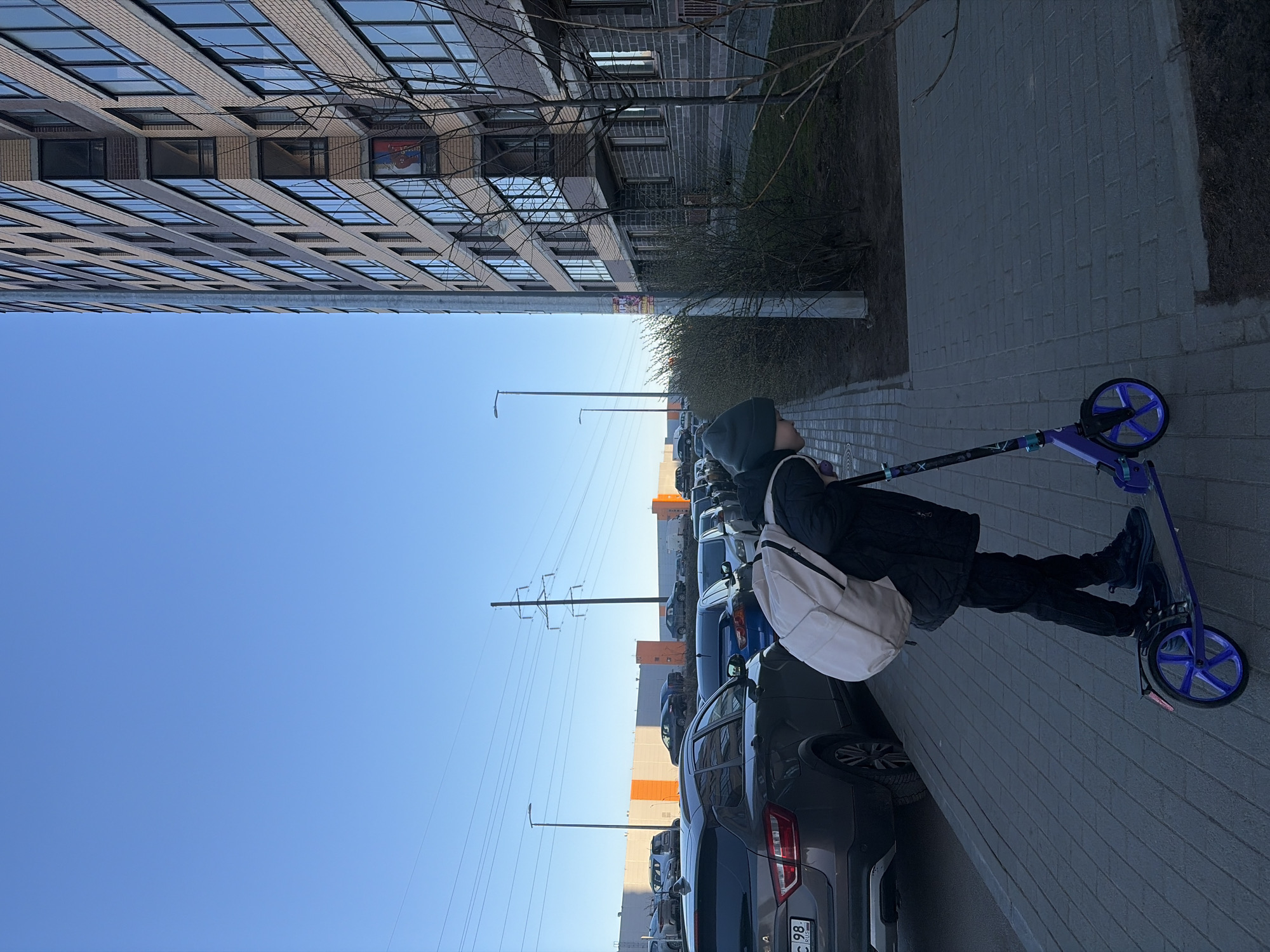 A young individual with a hood is standing on a purple scooter. The scooter is on a brick walkway parallel to parked cars. A tall building is in the background on the left. Overhead electrical power lines and streetlights are visible between the building and the cars. The license plate on a gray car closest to the foreground reads "C 98". The location is likely Saint Petersburg, Russia based on the license plate.