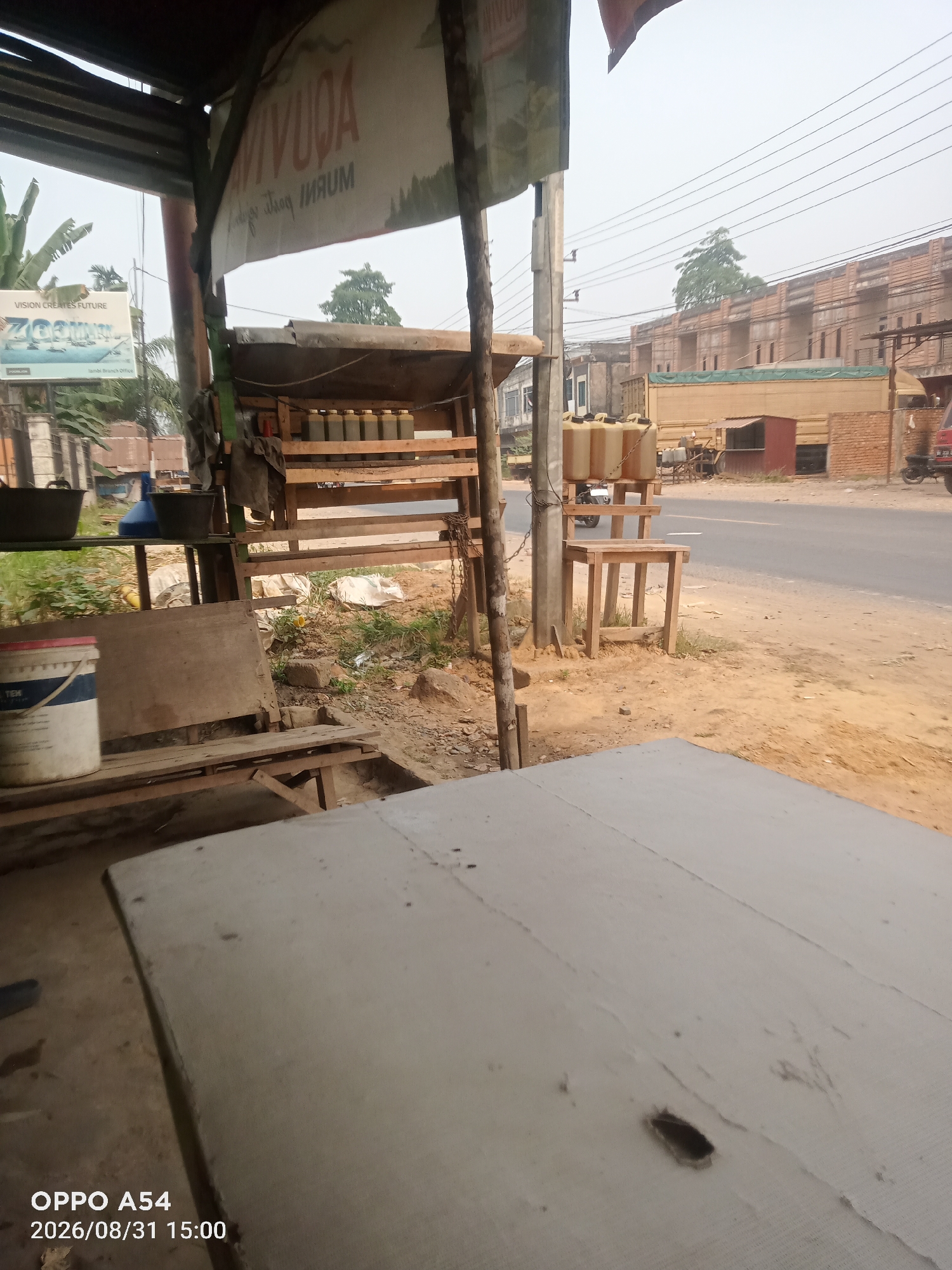 The image depicts an outdoor scene in Jambi, Indonesia, likely on the outskirts of the city or along a less developed road. In the foreground, a rough, light-colored table or surface occupies the lower portion of the frame. To the left, partially visible is a wooden bench. The middle ground features a makeshift wooden stall or structure, serving as a display and storage area. On one shelf of this stall are several rows of identical, light-colored bottles with yellow caps, possibly containing liquids. Below this, a white bucket with blue and red markings is visible.
Behind the stall, a dirt path or ground is visible, with scattered debris. Further back, across a paved road, are buildings under construction, characterized by their brick facades and scaffolding. The road itself is visible, with power lines strung overhead. A large truck with a green tarp is parked on the side of the road, and a motorcycle is parked near the stall. The sky is overcast, suggesting a cloudy day. The overall impression is one of a simple roadside vendor or workshop, with an urban backdrop of ongoing construction. The timestamp "OPPO A54 2026/08/31 15:00" is overlaid on the bottom left of the image.