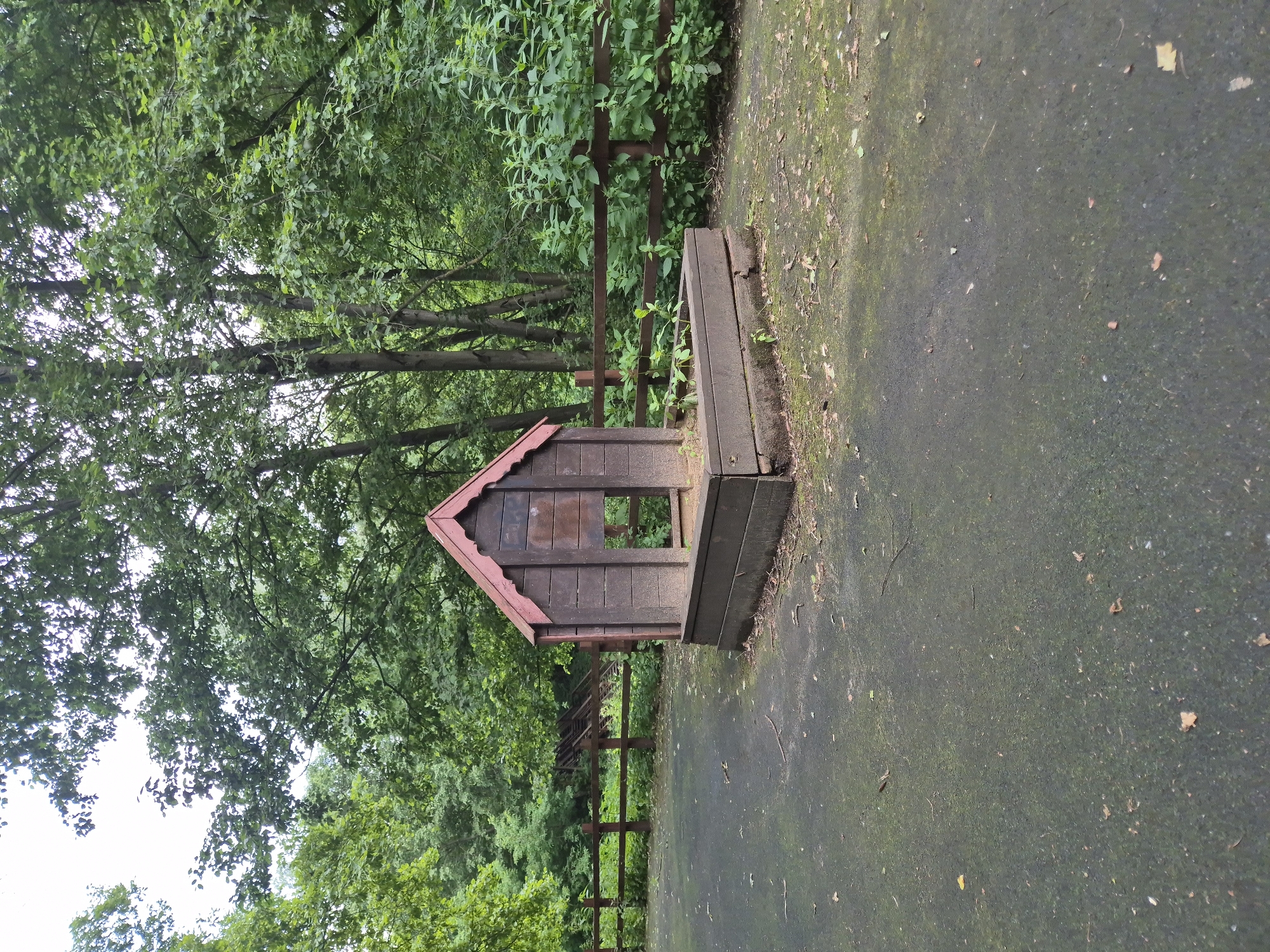 The image depicts a small, weathered wooden playhouse with a red roof, set on a dark, mossy paved surface. The playhouse is open in the front, revealing an empty interior. It is situated next to a wooden fence that encloses a densely vegetated area with tall trees and lush green foliage. The trees appear to be deciduous, with a full canopy of leaves, suggesting a warm season, likely summer or late spring. The overall scene is one of an outdoor play area or a section of a park or garden. There is no visible text in the image. The lighting suggests it is daytime, with diffused light indicating it might be overcast or the sun is high in the sky. The setting appears natural and somewhat rustic, with fallen leaves scattered on the paved area and around the playhouse. The image's orientation is rotated 90 degrees counter-clockwise.