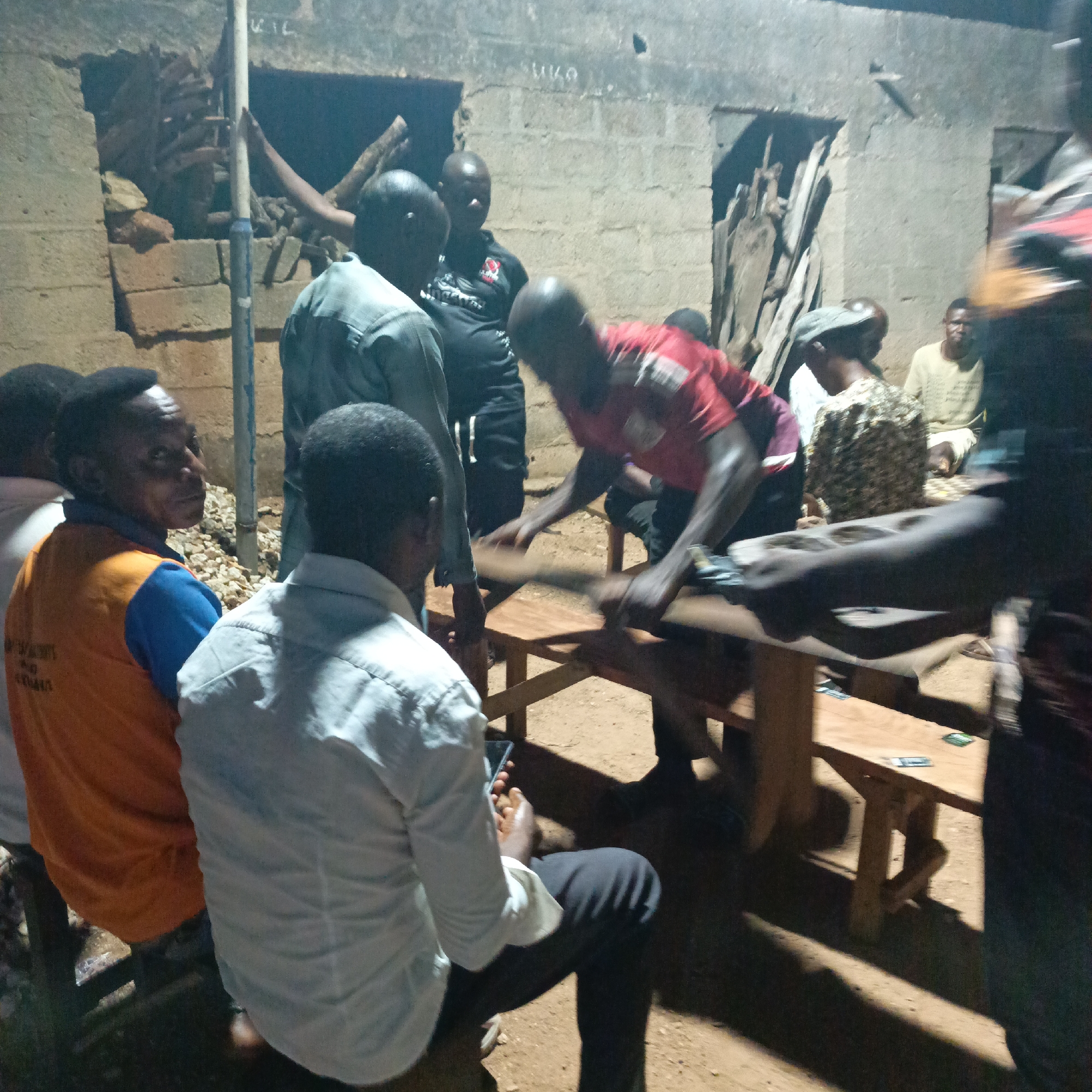 A group of people are gathered outside what appears to be a residential building in Akwanga, Nigeria. Some individuals are seated around a wooden table, while others stand. One person is actively engaged in playing a board game on the table. Some cards are visible on the table's surface. The building has a partially collapsed wall and a makeshift door. The setting appears to be lit by artificial light. One man is looking at his phone.