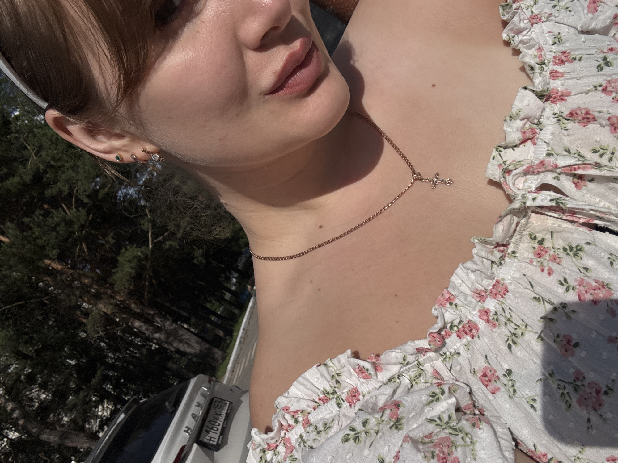 The image shows a close-up of a young woman's neck and shoulder, wearing a white floral print top with ruffled edges. A thin gold chain with a small cross pendant rests on her collarbone. Her skin is lightly tanned, and a small mole is visible on her neck. The background is blurred, showing dark green trees, suggesting an outdoor setting. A white car with a license plate that reads "H 160XX 159" is partially visible in the lower left corner. The lighting suggests it is daytime, possibly late afternoon, with dappled sunlight. The overall mood is casual and serene. The image appears to have been taken in Chaikovsky, Russia.