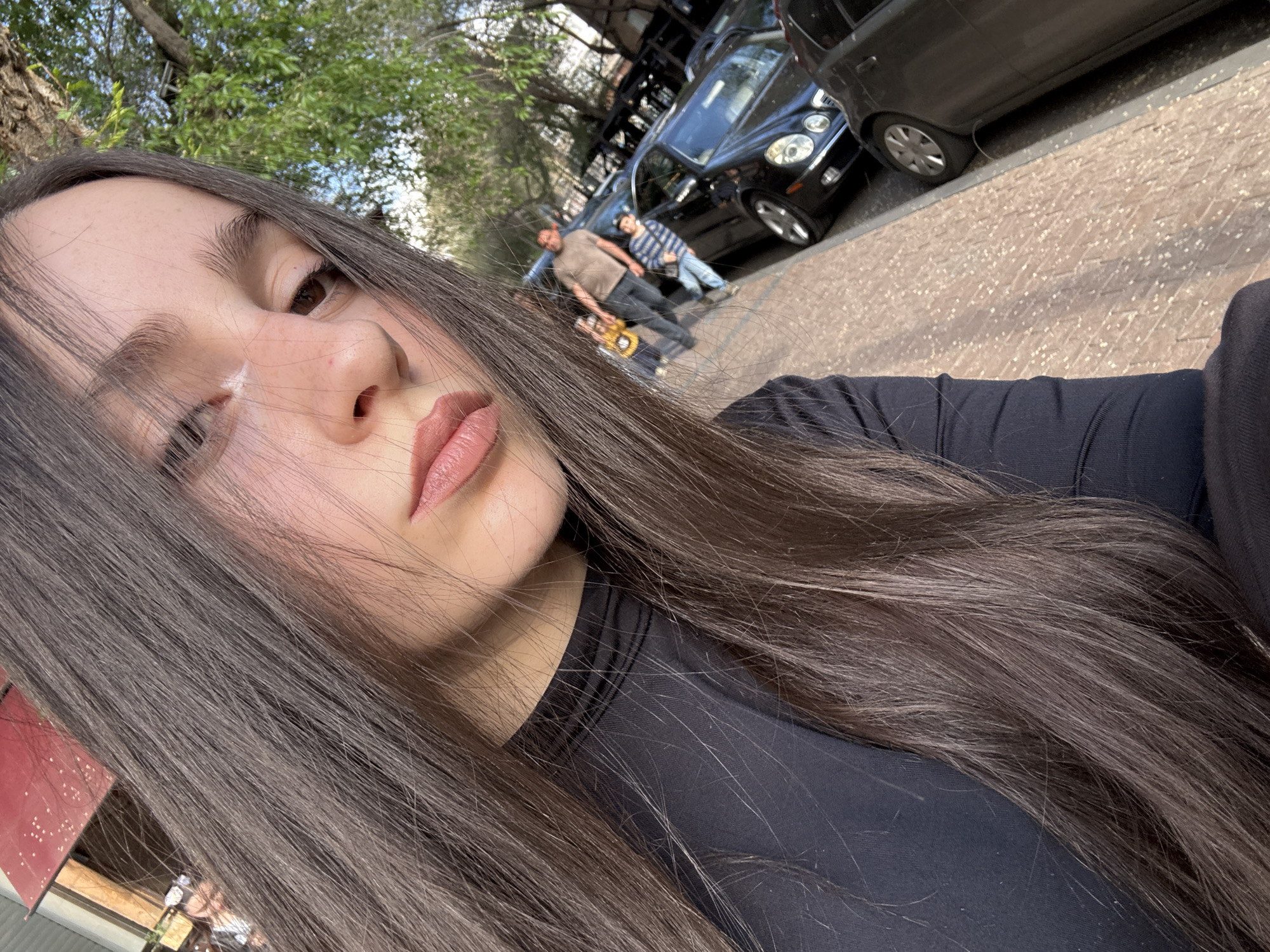 A woman with long, dark brown hair takes a selfie outdoors. Her face, seen from a close-up, tilted angle, occupies the left side of the frame, with her long hair cascading across her shoulder and chest, covering much of the bottom and left. She is wearing a black long-sleeved top and has a neutral expression with slightly parted lips showing muted pink lipstick.
In the background, out of focus, an urban street scene unfolds. Two individuals, an adult male in a light-colored t-shirt holding a yellow object (possibly a skateboard or scooter) and a child in a striped shirt, are standing on a paved sidewalk. Several dark-colored cars are parked along the curb, including a black luxury car with distinct front-end styling. Lush green foliage from trees is visible in the upper background, suggesting an outdoor, possibly treelined street. The ground is paved with light-colored bricks or cobblestones.
The lighting indicates it is daytime, with bright, possibly sunny, weather. The overall impression is a casual street scene in what is known to be Yerevan, Armenia. No discernible text is visible in the image.