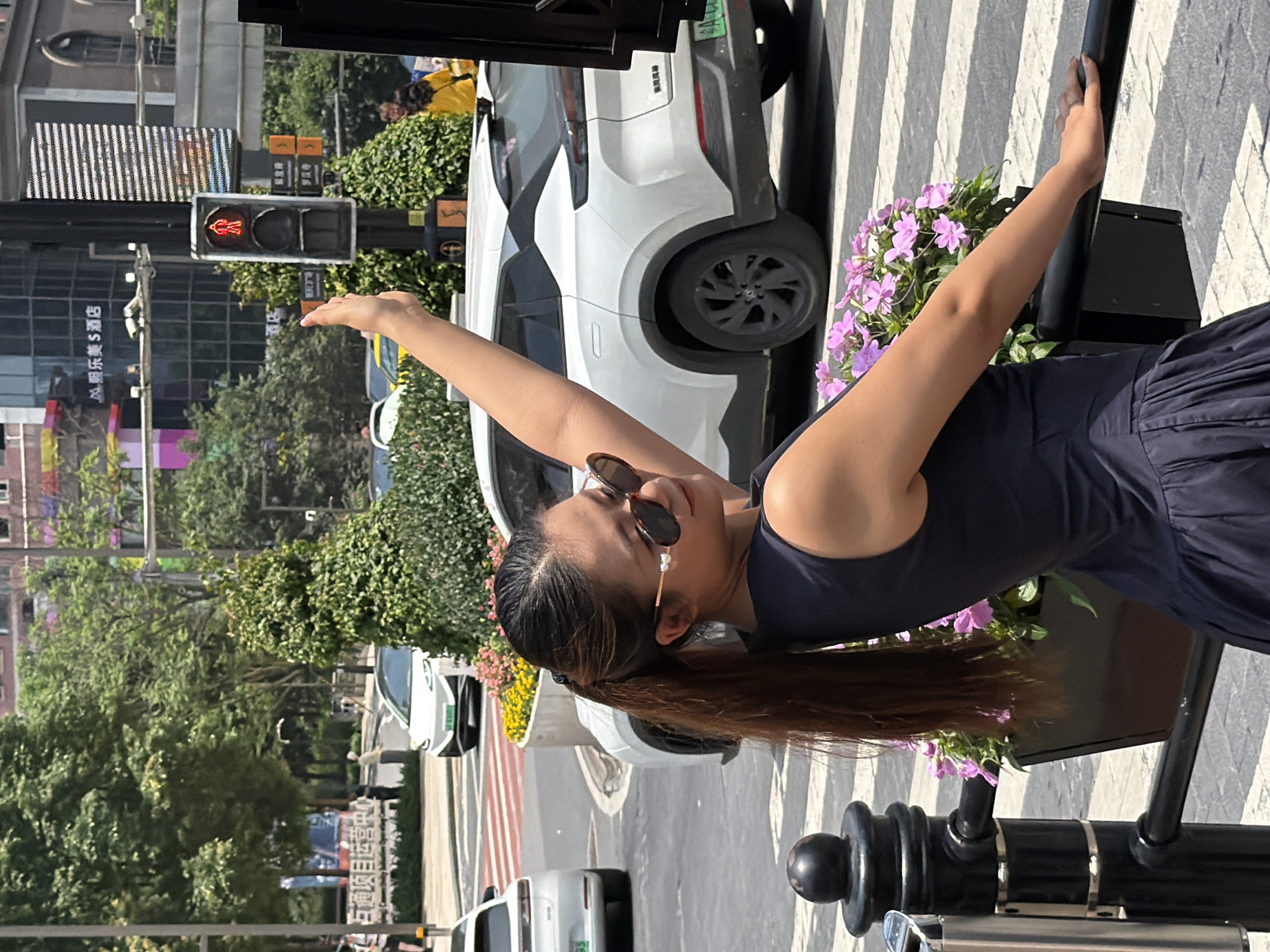 In Yuzhong District, China, a woman with long, dark hair pulled into a ponytail, wearing sunglasses and a dark sleeveless dress, stands on a street. She is leaning on a black railing, with a planter box of pink flowers to her left. Her right arm is raised, with her hand extended upwards. Behind her, a white SUV is stopped at a crosswalk, and traffic lights are visible. The scene appears to be a sunny day in an urban environment.