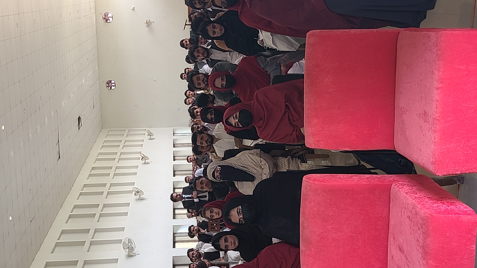A medium indoor shot shows a large gathering of people in an auditorium or hall, presumably in Quetta City Tehsil, Pakistan. A number of the individuals are men, a large portion wearing traditional attire. Many of the women in the foreground are wearing headscarves, some in black or red. Some of the women also wear a face mask. Many of the men have beards. A man in a suit can be seen. The walls are white and feature an ornate design with what appear to be decorative fan fixtures and overhead lighting. The ceiling is high with embedded lights. The foreground includes several large, stacked pink cubes or cushions. The overall scene suggests a lecture, meeting, or formal gathering.
