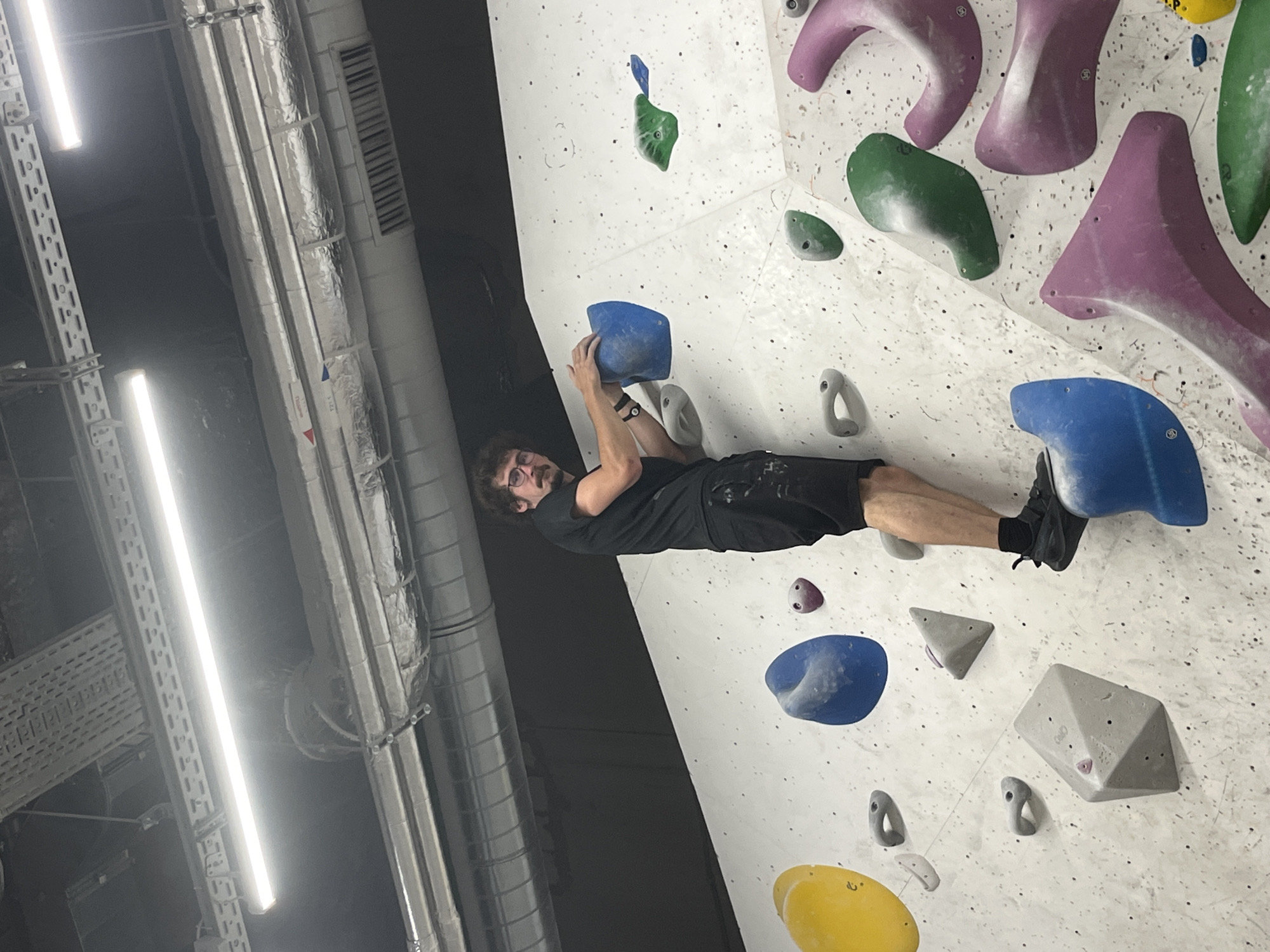 A man with dark, curly hair and a mustache, wearing glasses, a black t-shirt, and black shorts, is climbing a white bouldering wall. He is positioned in the center of the frame, looking towards the camera. His left hand is resting on a large blue climbing hold, and his right hand is reaching for another blue hold. His left foot is on a large grey triangular hold, and his right foot is on a large blue hold. The bouldering wall is covered with various colorful climbing holds of different shapes and sizes, including yellow, blue, grey, green, and pink. Above the wall, industrial pipes and fluorescent lighting are visible on the ceiling. The scene appears to be an indoor climbing gym. The provided location of Khimki, Russia, is a city in the Moscow Oblast.