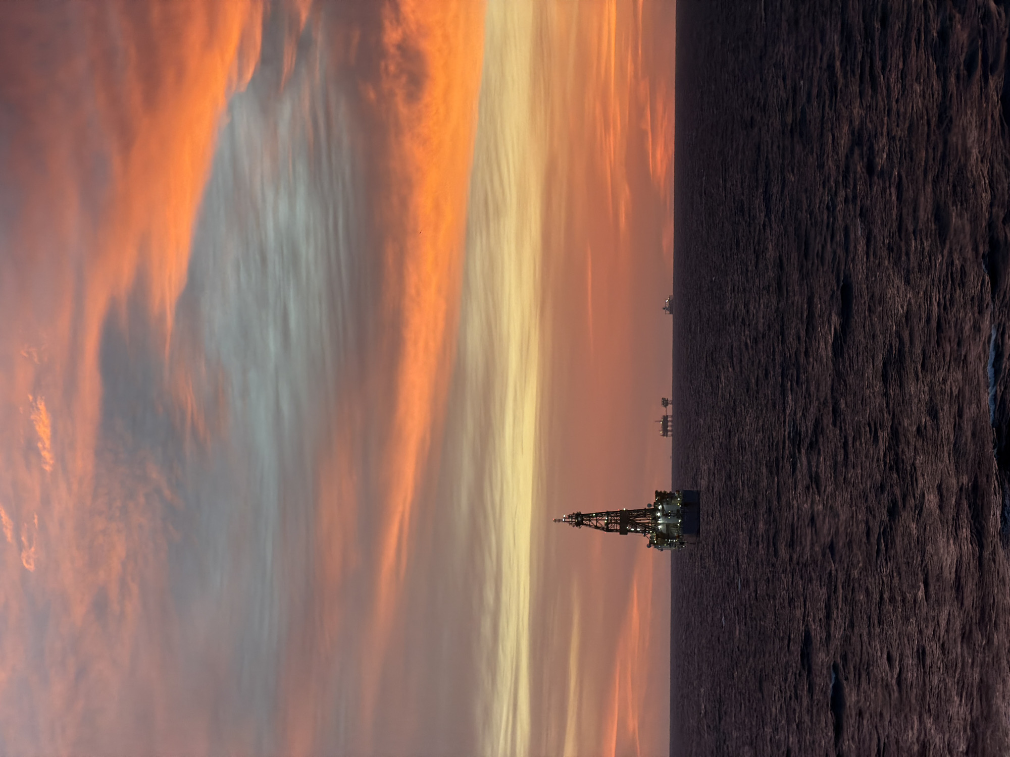 The image depicts an offshore oil rig at sea during twilight or sunrise. The sky is a mixture of orange, yellow, and grey striated clouds. The sea is dark, contrasting with the lit-up rig. Two additional oil rigs are visible in the distance.