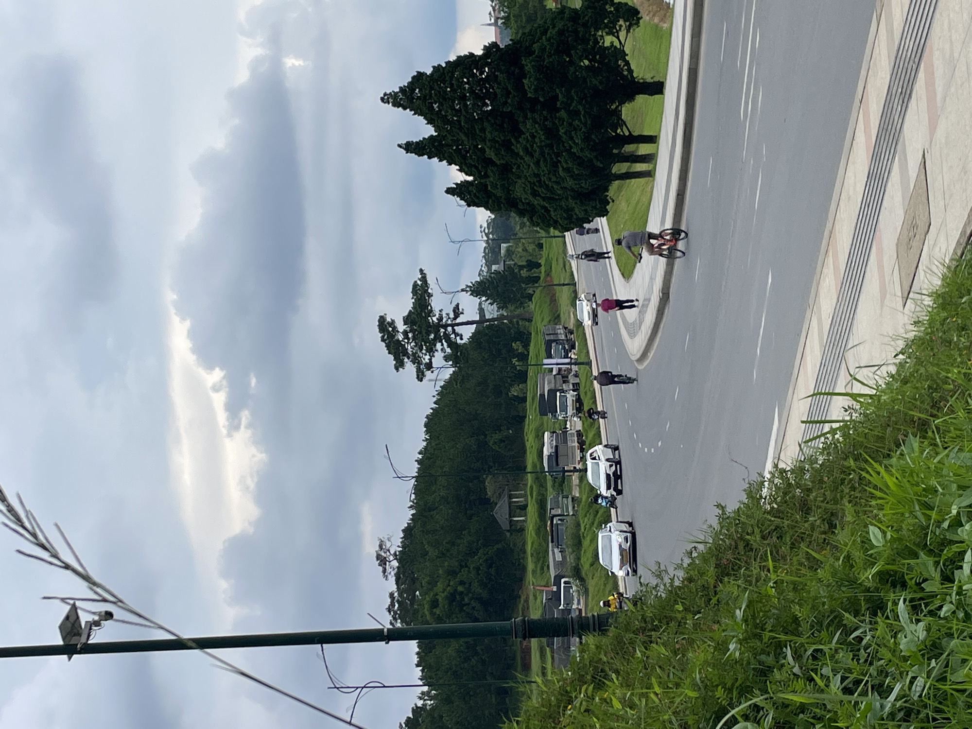 The image depicts a cloudy day in Da Lat, Vietnam, on a winding asphalt road lined with grass and trees. On the left side of the road, a tall street lamp with a solar panel is visible. Several cars and motorcycles are driving on the road. A person is riding a bicycle on the right side of the road, and another person is walking on the sidewalk. In the background, a dense forest covers a hill, with a small pavilion visible among the trees. Trucks are parked on the side of the road in the distance. The overall scene suggests a residential or suburban area of Da Lat with ongoing traffic and recreational activities.