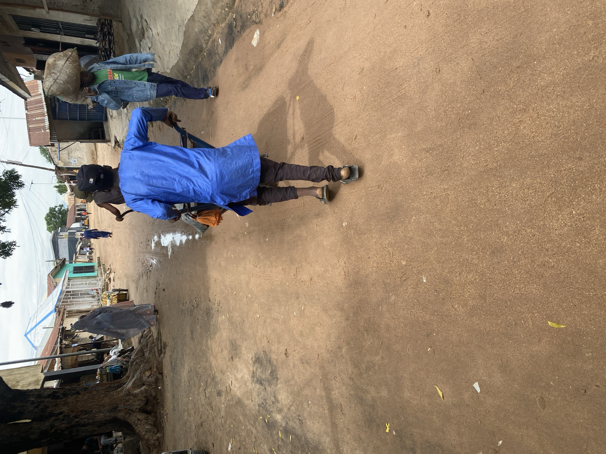 The image depicts a street scene in Jalingo, Nigeria, with two men as the primary subjects. The man in the foreground is wearing a blue tunic and dark pants, pushing a cart. He has a dark cap on his head. The man in the background is carrying a large sack of goods on his head, wearing a green t-shirt and a denim jacket. He is walking away from the viewer.
The setting is a dirt road with buildings and structures lining the sides. Some of the buildings appear to be shops or homes, with corrugated metal roofs and simple construction. There are some scattered debris and foliage on the ground, indicating a possibly dusty or rural environment. A large tree with exposed roots is visible on the left side of the image. The lighting suggests it is daytime, and the sky is overcast, hinting at potentially cloudy or humid weather.
The activity taking place is the movement of people and goods through the street, suggesting a busy marketplace or thoroughfare. The man in the foreground is engaged in transporting something via a cart, while the man in the background is carrying a heavy load, implying labor or commerce. No text is clearly visible in the image. The overall impression is that of a typical day in a Nigerian town, with people going about their daily activities.