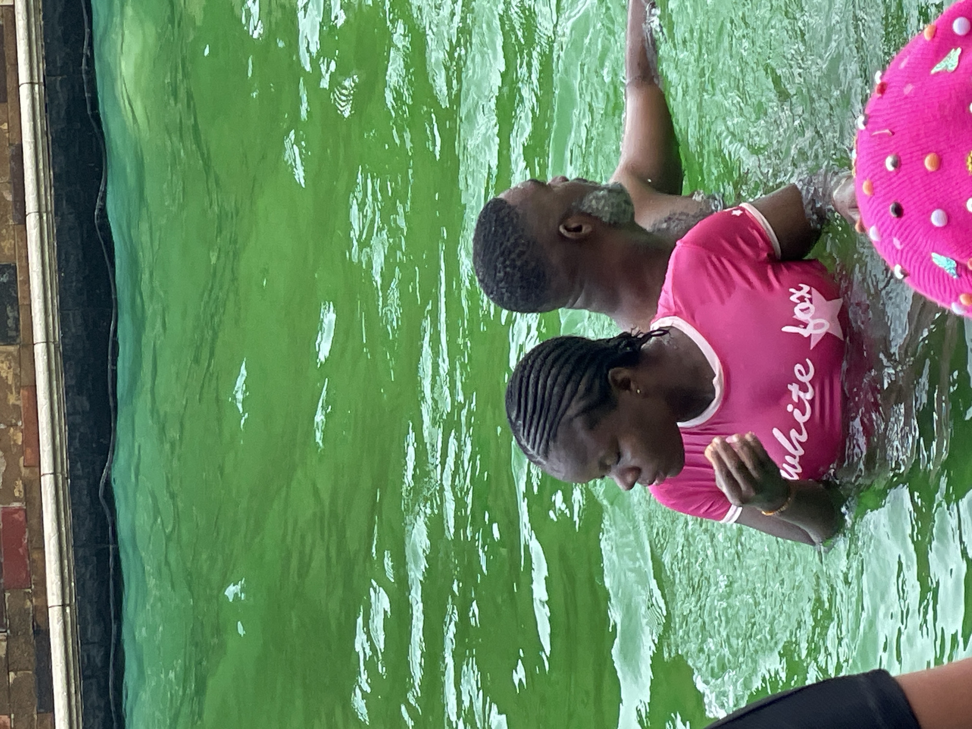 Two individuals are submerged in a bright green swimming pool. In the foreground, a Black woman with braided hair wears a pink t-shirt with "white fox" printed in white cursive script. She is looking down and holding something to her mouth. Behind her, a Black man with graying facial hair and a tattoo on his arm is partially visible, looking to his right. The pool water is choppy and reflects light. In the background, the edge of the pool is visible, constructed with brick and white coping.
