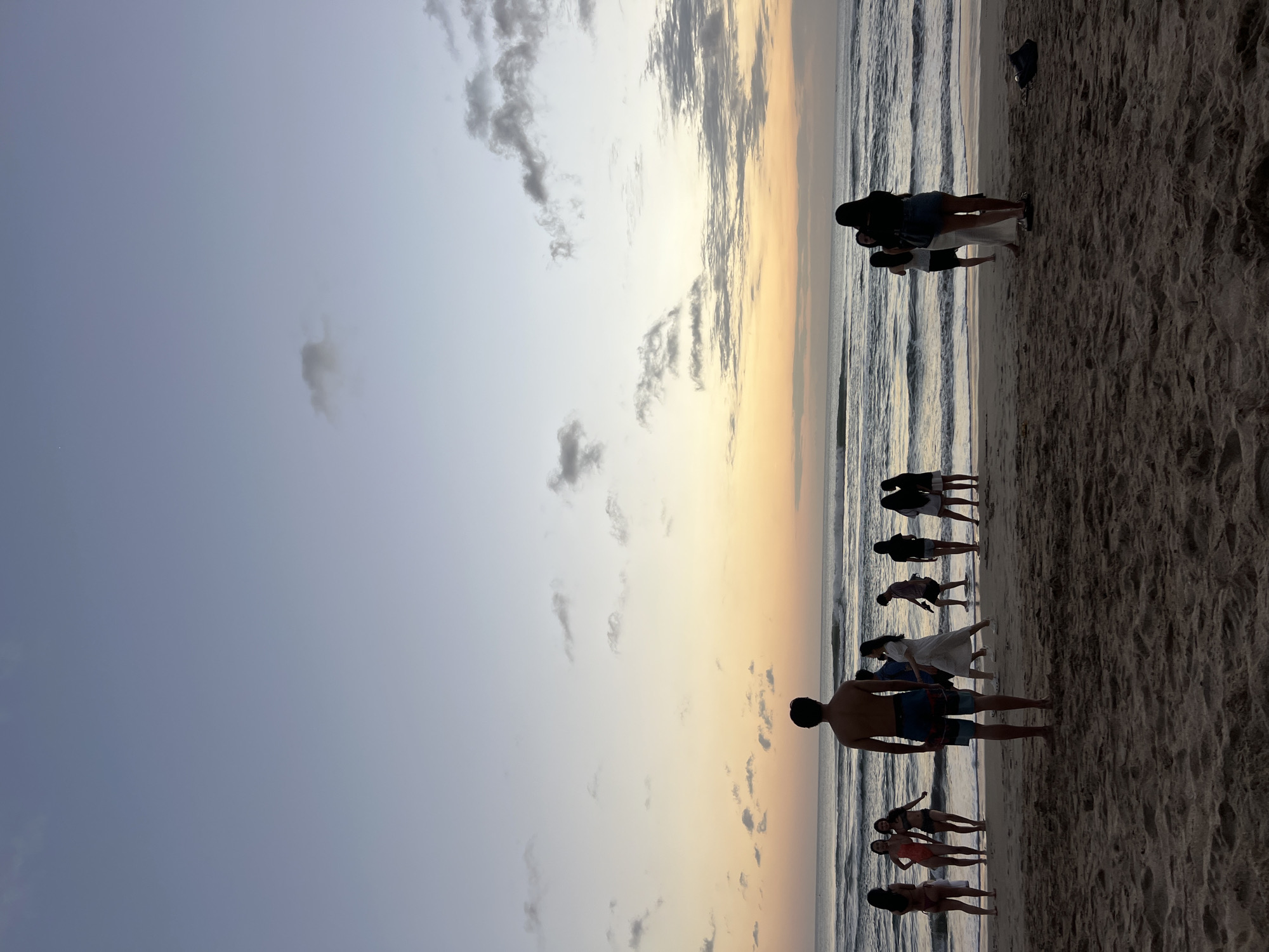 In Kuta, Indonesia, a group of approximately ten people stands on a sandy beach at dusk. The sky is a gradient of soft blues, oranges, and yellows, with scattered clouds. The ocean waves are gentle, and the tide is out, revealing a wet sand area near the water. Most of the people are facing the ocean, with some standing in small groups and others individually. A man in swim trunks stands in the foreground, facing away from the camera. Several women in swimwear are gathered on the left, and other individuals are positioned further down the beach. The scene is serene, with the warm evening light casting long shadows on the sand.