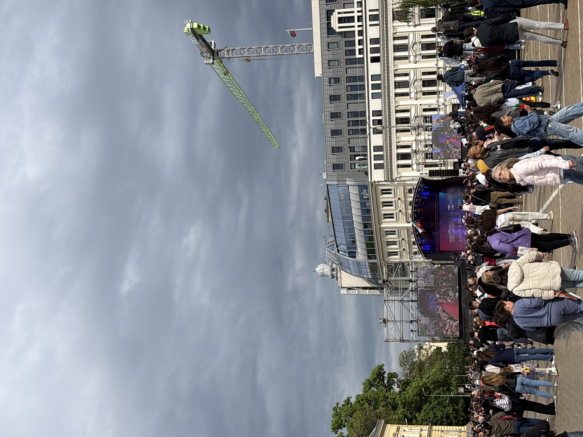 The image captures a large crowd gathered in Sofia, Bulgaria, likely at a public event or celebration. The crowd, composed of people of various ages and ethnicities, is facing a stage where a large screen displays the Eurovision Song Contest. Many people are holding Bulgarian flags. The event is set against a backdrop of tall buildings, including a modern glass-faced structure and a traditional stone building. A construction crane is visible in the background. The sky is overcast and grey.