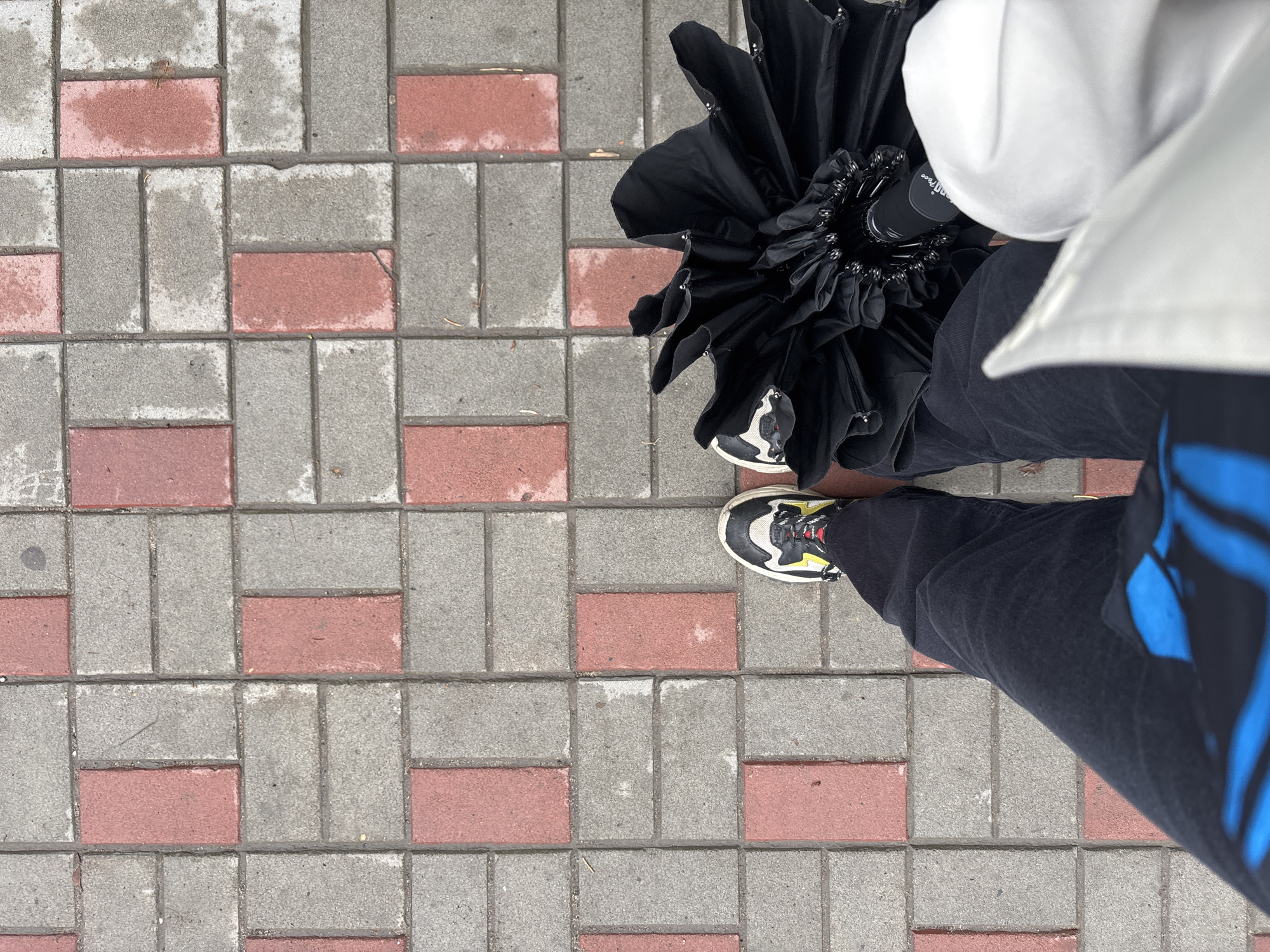 The image is a top-down view of a person standing on a sidewalk in Nizhny Novgorod, Russia. The sidewalk is paved with interlocking rectangular bricks, alternating between gray and red. The person is wearing black pants with a blue design and black and white sneakers with yellow laces. They are holding a partially opened black umbrella. A white garment, possibly a coat or bag, is also visible. The ground appears slightly damp, suggesting recent rain.