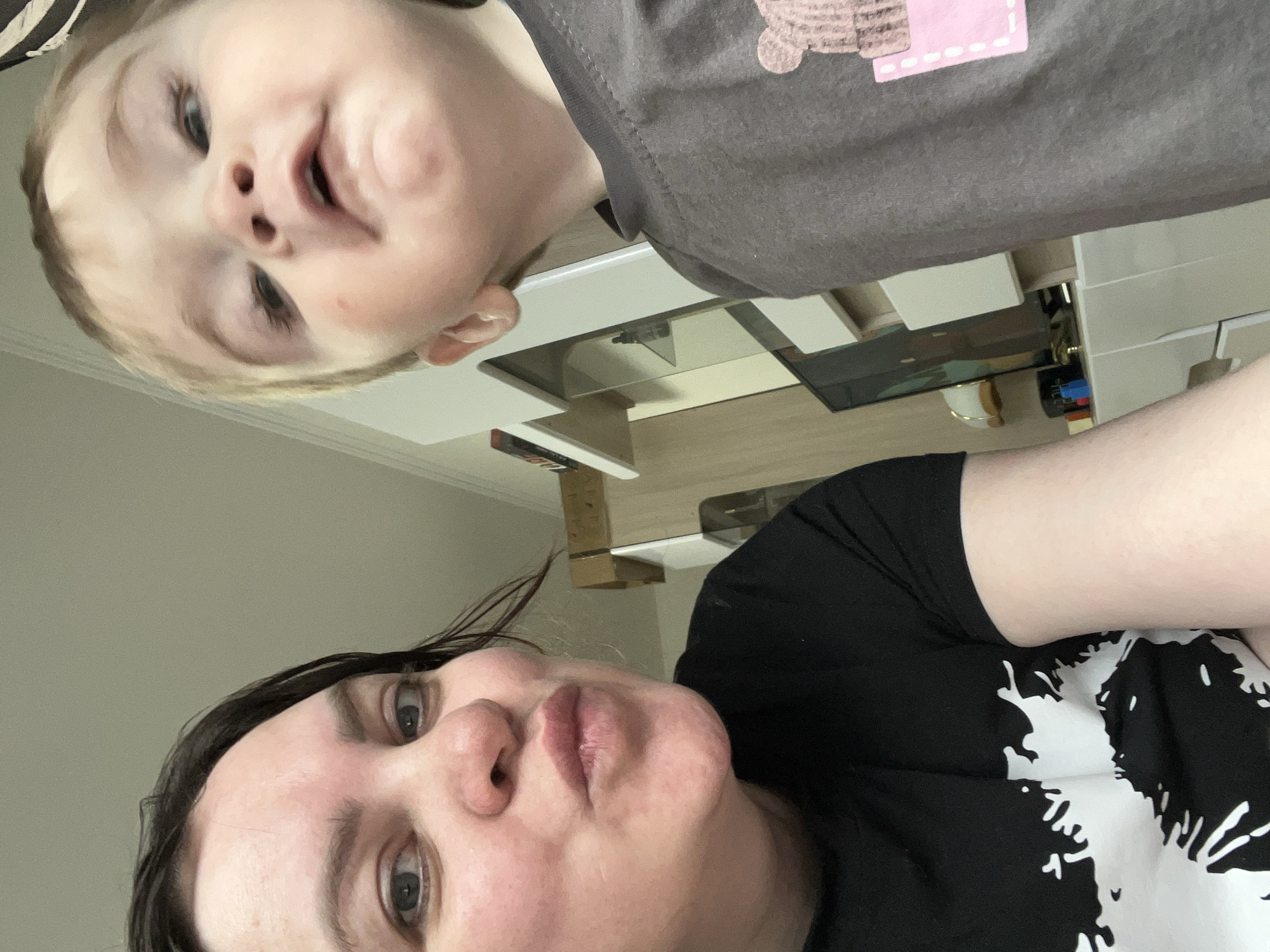 A rotated, low-angle indoor photograph captures a woman and a young child looking directly into the camera. The image is oriented such that the woman is positioned on the lower right and the child on the upper left, both appearing to look down towards the camera.
The woman has dark hair and light skin, displaying a neutral expression. She is wearing a black t-shirt with a large white, abstract design. The child, with light-colored hair and fair skin, has a playful or slightly smirking expression. The child is dressed in a dark grey t-shirt featuring a pink rectangular patch with dashed outlines and a brown textured shape on the shoulder.
The background suggests a home interior, likely a living room or kitchen area in Perm, Russia. A light-colored table or counter surface is visible in the middle ground, with a gold-colored cup resting on it. Behind the table, various furniture pieces or shelves can be seen, along with a white ceiling fixture near the woman's head. The lighting indicates it is daytime, and the photograph appears to be a casual selfie or portrait taken from a low vantage point.
