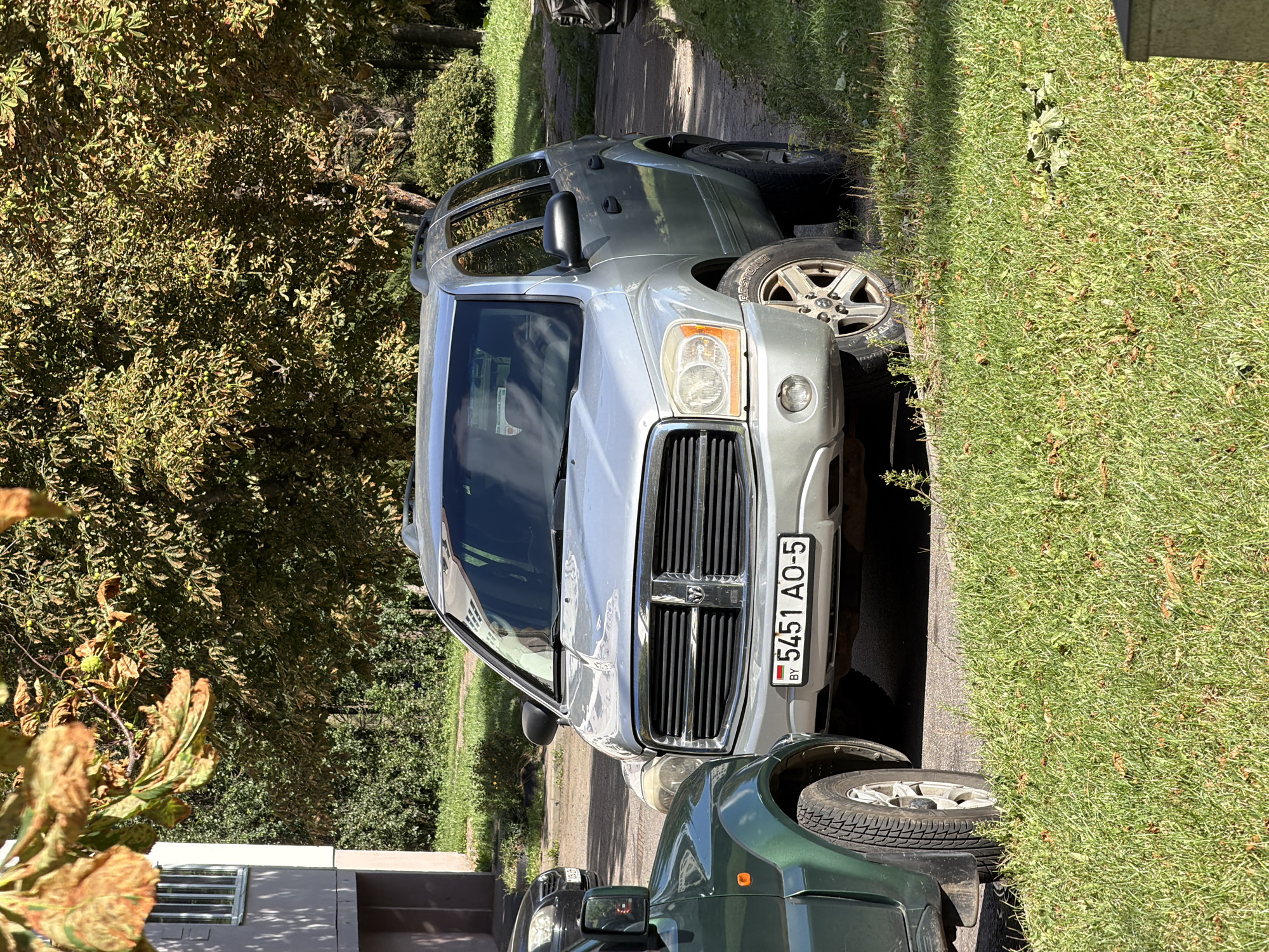 In Minsk, Belarus, on a sunny day, a silver Dodge Durango SUV is parked on the right side of a paved area, partially obscured by a green vehicle on the left and a building on the far left. The Durango's license plate reads "BY 5451 AO-5". Fallen leaves are scattered on the grass and ground around the vehicles, indicating it might be autumn. Trees with green and yellowing leaves, and some horse chestnuts, overhang the scene. A portion of a building with a window is visible to the left, and more greenery and trees form the background. There are no people visible in the image.