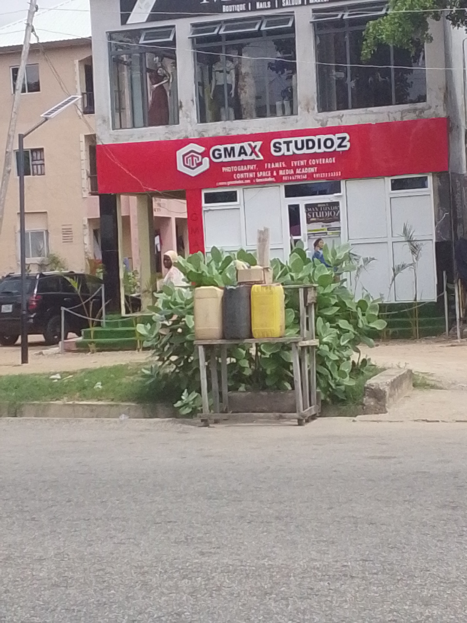 The image captures a street-level view of a commercial building in Jalingo, Nigeria, with a prominent red signboard displaying "GMAX STUDIOZ" and listing services like "PHOTOGRAPHY, FRAMES, EVENT COVERAGE, CONTENT SPACE & MEDIA ACADEMY". A black SUV is parked to the left of the building. In front of the building, a display rack holds three large containers: one yellow, one light brown, and one dark gray. Lush green foliage surrounds the display rack. A person wearing a light-colored head covering and white garment stands to the right of the SUV, near the building entrance. The building itself appears to have large windows, some of which display mannequins. The scene is situated on a paved street with a sidewalk.