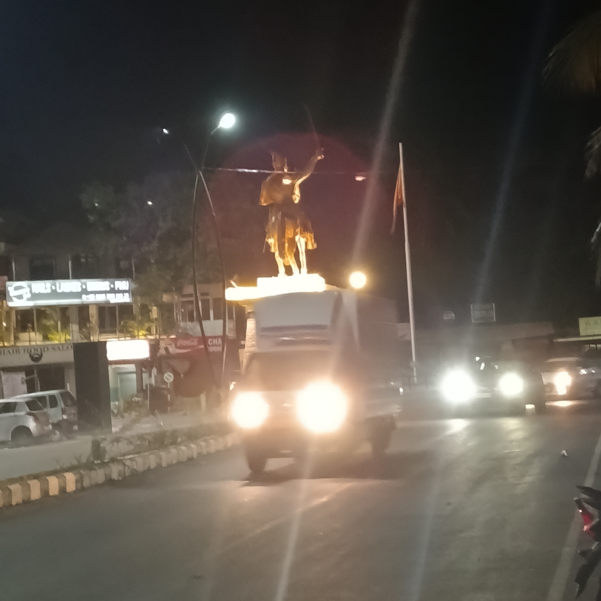 At night, a vehicle with bright headlights drives on a street in Oxel, India, obscuring the base of a statue of a man on a horse holding a sword. Behind the statue, buildings line the street, one with a sign reading "NAILS LADIES BROWS PERM." A flag is visible to the right of the statue. Other vehicles are on the street, and the street is bordered by a low wall.