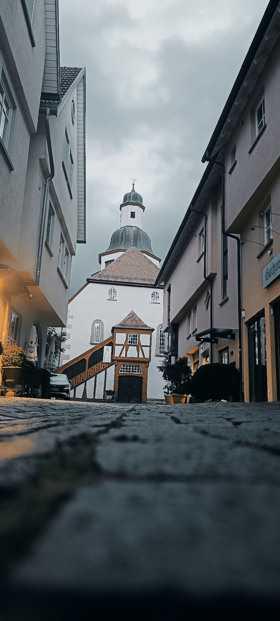 The image depicts a narrow street in the historic center of Winnenden, Germany, featuring traditional European architecture. The perspective is low, emphasizing the cobblestone street and the surrounding buildings. On the left, a light-colored building with windows and a gutter system dominates the foreground. In the center, a striking white building with a bell tower and a pitched roof stands out. Attached to this building is a distinctive half-timbered structure with an external staircase leading up to its entrance. The staircase is painted in warm, natural wood tones with dark accents. The entrance to the half-timbered section features a large, dark wooden door. On the right, another building, painted in a pale peach hue, shows windows and a dark awning over a shop entrance. The sky is overcast and grey, contributing to a muted color palette. Several potted plants are visible, adding touches of green to the scene. A dark-colored car is parked on the left side of the street, and a table and chairs are set up on the left, suggesting an outdoor seating area. The overall impression is one of historical charm and a quiet urban setting.