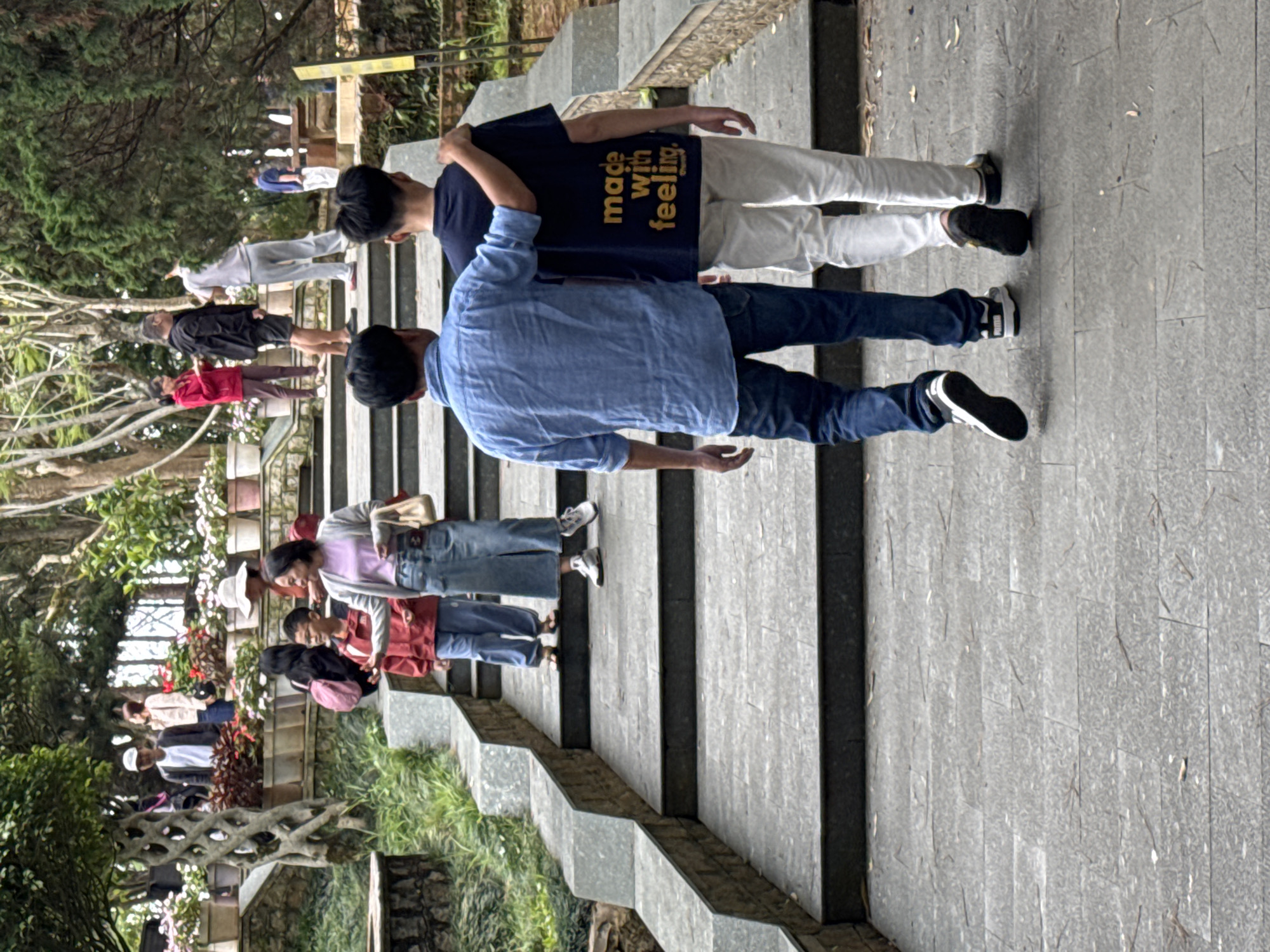 In Da Lat, Vietnam, two young men are walking up a stone staircase in a park-like setting. The man on the left, wearing a blue long-sleeved shirt and blue jeans, has his arm around the shoulders of the man on the right. The man on the right wears a dark t-shirt with yellow text that reads "made with feeling" and white pants. They are walking away from the camera. Other people are visible further up the stairs and in the background, including a woman in a red jacket, a man in a black jacket, and several others. The staircase is flanked by greenery and trees. Potted plants are visible on ledges beside the stairs. The ground is paved with grey stone tiles.
