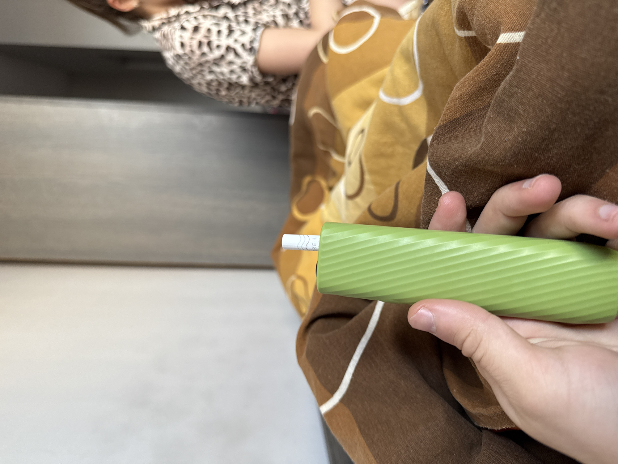 A person is holding a green, textured object in their hand. The object has a white tip. The person is sitting on a brown and tan patterned blanket. Part of a child wearing a patterned shirt is visible in the upper left of the frame. A light gray surface is also visible in the background on the left side of the frame. The location is unknown.