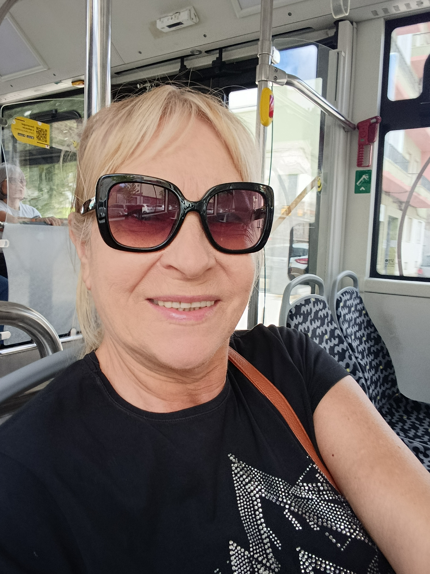 A woman with blonde hair, wearing sunglasses and a black t-shirt, is taking a selfie inside a bus. She is smiling, and her face is visible from the shoulders up. A brown strap from a bag is visible across her chest. In the background, there are seats with a patterned fabric, windows showing a city view with buildings and trees, and various fixtures of the bus interior, such as handrails and buttons. A QR code is visible on a yellow sticker on a protective barrier. The scene appears to be in the city of Setúbal, Portugal.