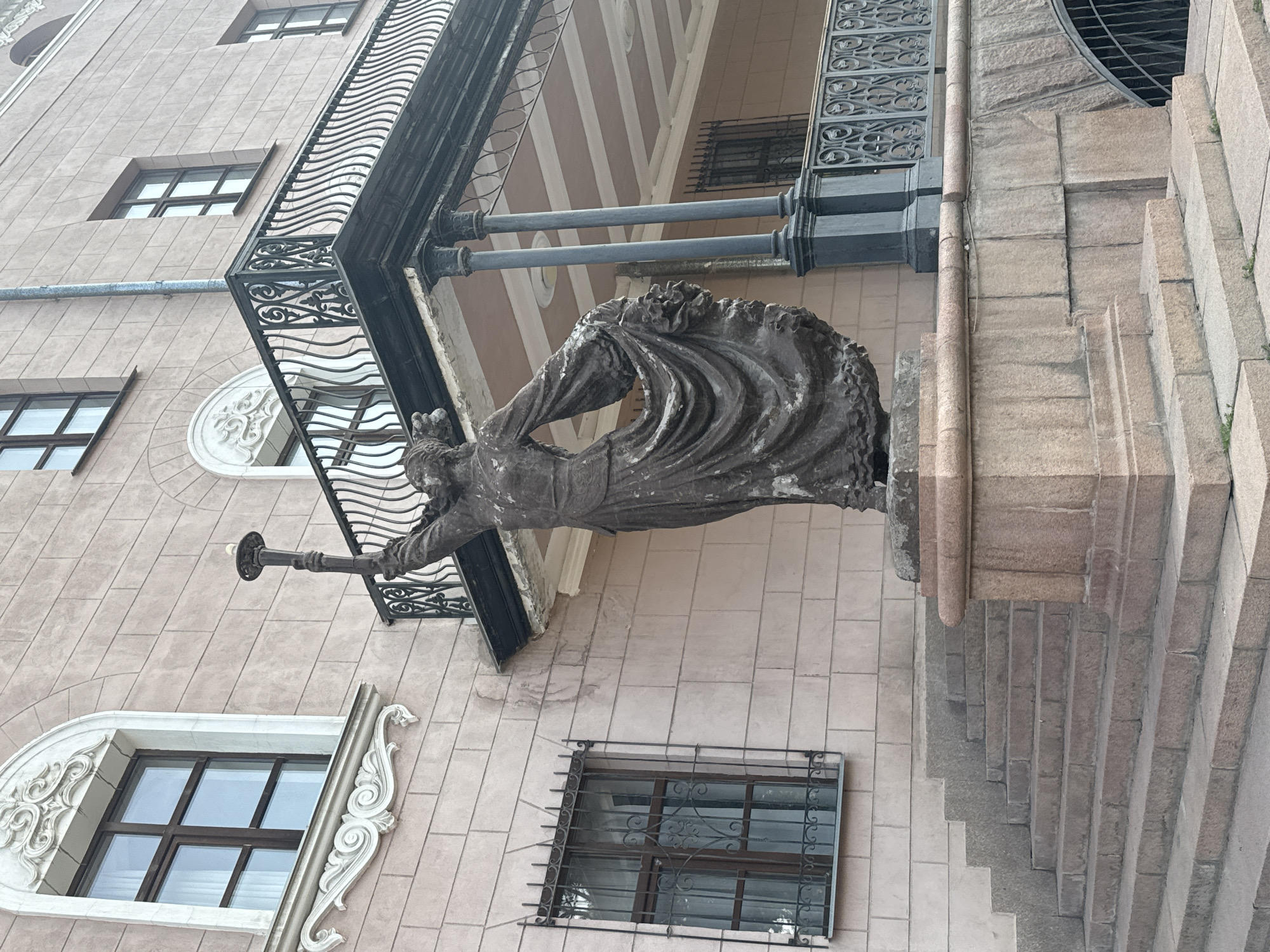 The image shows a bronze statue of a woman holding a torch aloft, with her dress dramatically billowing. The statue stands on a stone pedestal at the bottom of a wide, multi-tiered stone staircase. Behind the statue and to the right, a building with a pinkish facade is visible, featuring ornate windows and a black metal balcony with decorative scrollwork. The building also has a prominent drainpipe running vertically down the facade. The scene appears to be outdoors, with hints of greenery at the base of the stairs. This statue is located in Ala-Too Square, in Bishkek, Kyrgyzstan, and represents Liberty.