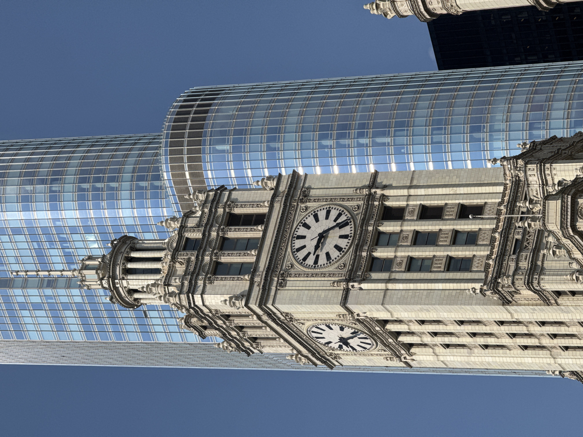 The image captures a close-up, low-angle view of the historic Wrigley Building clock tower in Chicago, Illinois, with modern glass skyscrapers in the background. The Wrigley Building, a white limestone structure, features two large, circular clocks with black hands and Roman numerals. The clock on the left shows approximately 4:48, and the one on the right shows approximately 4:49. Intricate architectural details adorn the tower, including cornices, decorative molding, and multiple rows of windows. Behind the Wrigley Building, a tall, slender skyscraper with a glass facade dominates the scene, its blue panes reflecting the clear sky. To the right, a curved glass building partially obstructs the view of another dark skyscraper. The sky is a deep, clear blue. The photograph is taken from a street-level perspective, emphasizing the verticality and scale of the architecture.