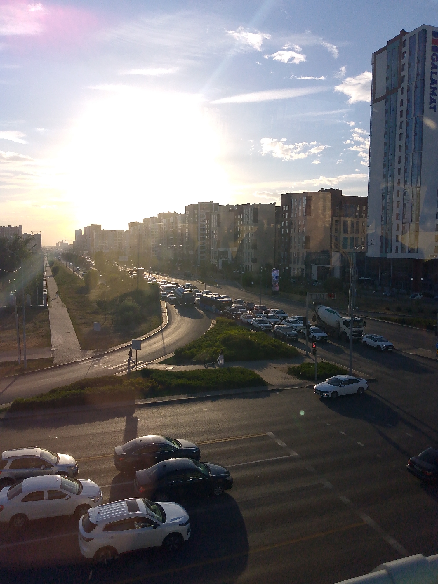 This is a vertical, eye-level, medium-range shot of a busy city street during sunset. The sun is low in the sky, casting a warm glow and creating long shadows. The sky is a mix of blue and white with some wispy clouds, and there are lens flares visible from the bright sun.
The scene depicts a multi-lane road with a significant amount of traffic. Several cars are visible, including white sedans, SUVs, and darker colored vehicles. There are also larger vehicles such as a cement truck and a bus. Some pedestrians are seen on the sidewalk and crossing the street at a marked crosswalk.
The setting is urban, characterized by several modern high-rise buildings lining the street. One building on the right has the word "GALAMAT" written vertically in large blue letters. The architecture suggests a contemporary city. The road is paved and marked with lane lines. There are also traffic lights visible, one of which appears to be red. Greenery, including trees and bushes, is present along the sides of the road and sidewalks.
The overall impression is one of a bustling urban environment at the end of the day, with the golden hour light adding a touch of warmth to the scene. The presence of numerous vehicles and pedestrians suggests normal daily activity. The location is unknown, but the modern architecture and the name on the building offer some clues.