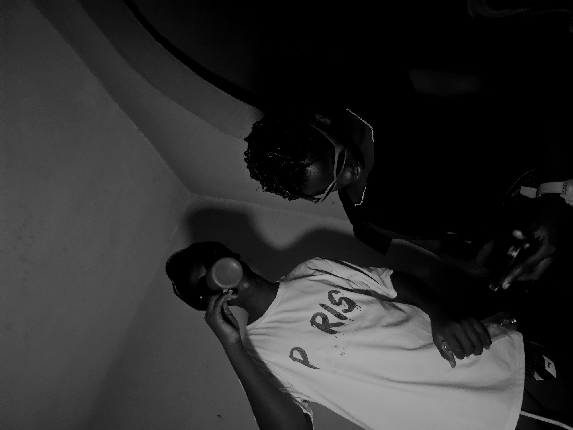 Two individuals are visible in the monochromatic image. One person, wearing a "PARIS" T-shirt and a cap, is holding a cup to their mouth. The other individual, standing above them, has braided hair, glasses, and is holding a glass bottle and a lighter. The setting appears to be indoors with plain walls and a dark background. The location is in Nkwelle, Nigeria.