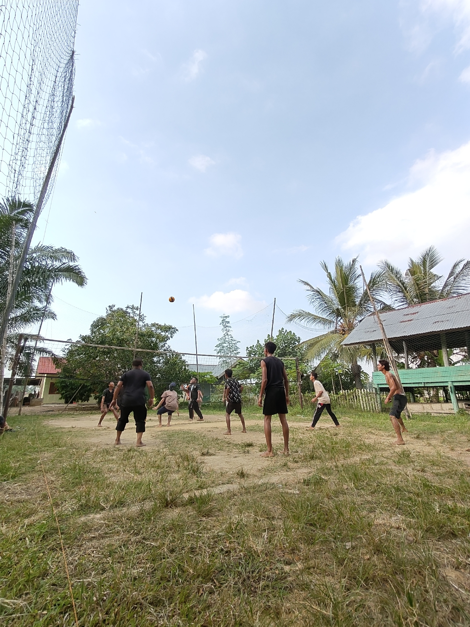A group of eight young men are playing volleyball on a dirt court in Ubitpayaitek, Indonesia. The sky is a light blue with a few scattered clouds. Palm trees and other foliage are visible in the background, along with a simple building with a corrugated metal roof. The players are dressed in casual attire, mostly shorts and t-shirts, and appear to be enjoying the game. One player in the center of the frame is jumping to hit the ball, which is in mid-air. The net is stretched across the court, and the poles supporting it are made of rough wood. The ground is a mix of dirt and dry grass. The scene suggests a casual, outdoor recreational activity in a tropical or subtropical environment.