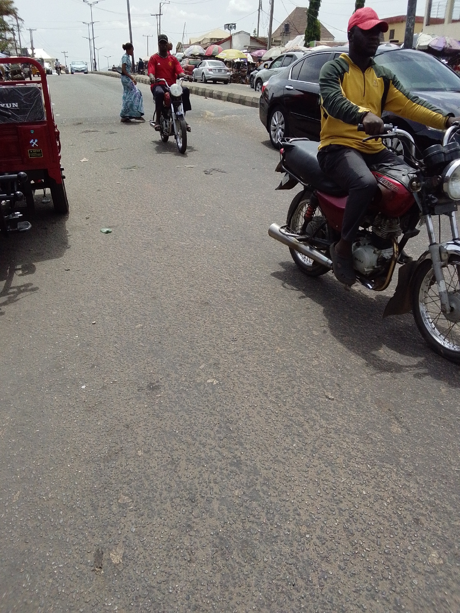 The image depicts a street scene in Lokoja, Nigeria. A person wearing a red cap and a yellow and green jacket is riding a red motorcycle in the right-hand side of the frame. Another person in a red shirt is riding a motorcycle behind them, and a woman in a blue patterned dress is walking along the side of the road in the background. A red tricycle with "YUN" printed on the back is partially visible on the left-hand side of the frame. Several cars are parked along the roadside, and a few buildings are visible in the background. The sky is overcast.
