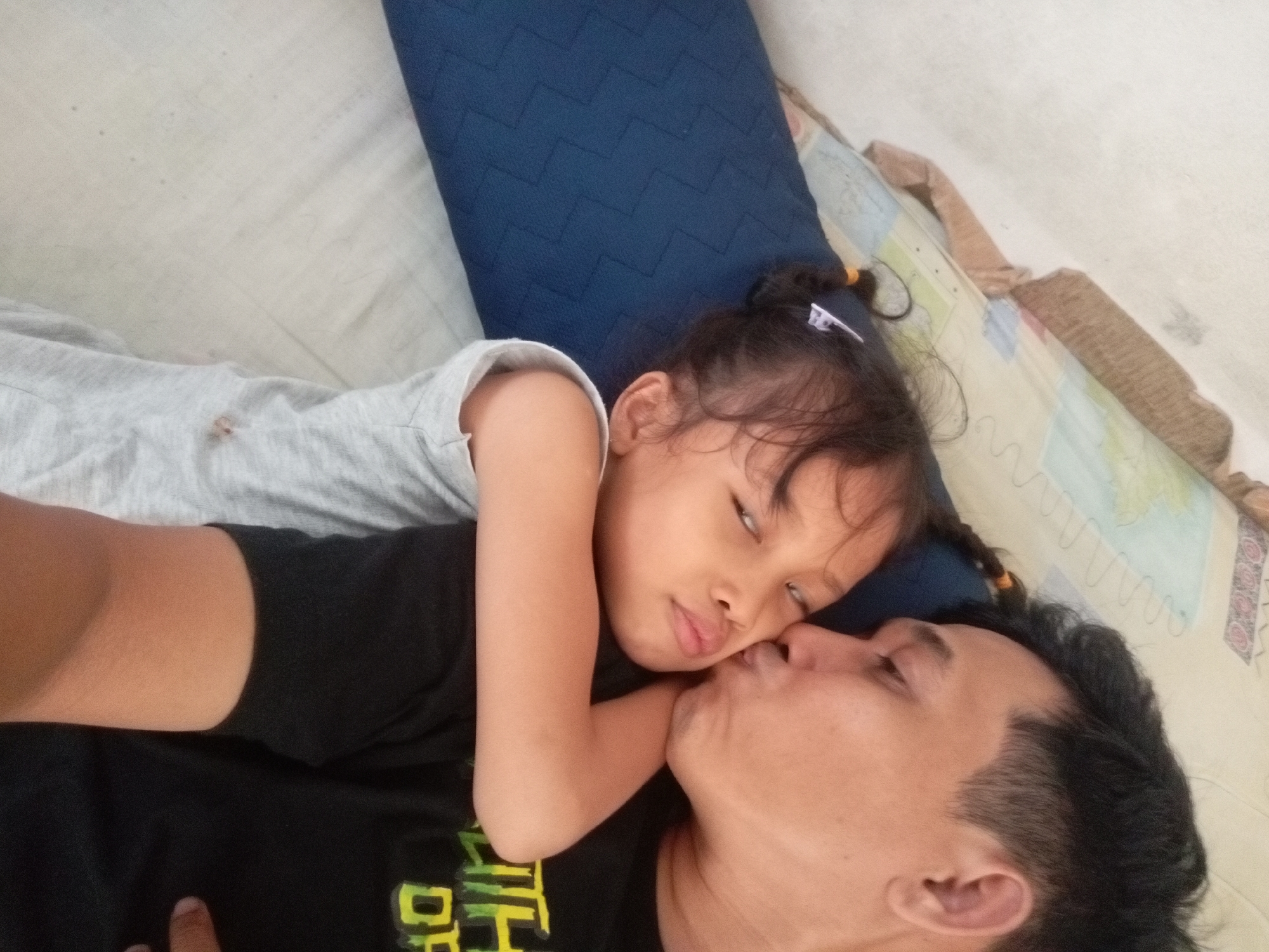 The image shows a father and daughter lying together on a bed. The father is kissing his daughter on the cheek. The daughter is leaning her head on her father's chest. They are both wearing casual clothing. The room appears to be a bedroom, with a pillow and bedding visible. The lighting suggests it is daytime. The father and daughter appear to be sharing a tender and loving moment. No visible text.