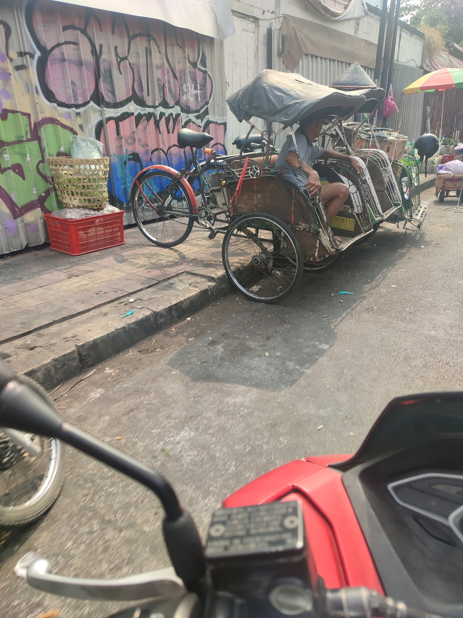 The image depicts a street scene in Yogyakarta, Indonesia, with a focus on a becak, a three-wheeled passenger pedicab. The becak is parked on the side of the road, which appears to be paved with asphalt. In the foreground, the handlebars and part of the red body of a motorcycle are visible, suggesting the photo was taken from a motorcycle's perspective.
Behind the becak, a corrugated metal wall is covered with colorful graffiti. To the left of the becak, a woven basket filled with goods sits atop a red plastic crate. The becak itself has a canopy providing shade and appears to be made of wood and metal. A person, dressed in a light blue t-shirt and shorts, is seated in the driver's position of the becak, looking to the side. Several other becaks are parked or in motion further down the street. An umbrella with red, yellow, and green panels is visible in the background, along with other stalls and structures that suggest a market or roadside commerce. The sunlight indicates it is daytime, with shadows cast on the road and sidewalk.