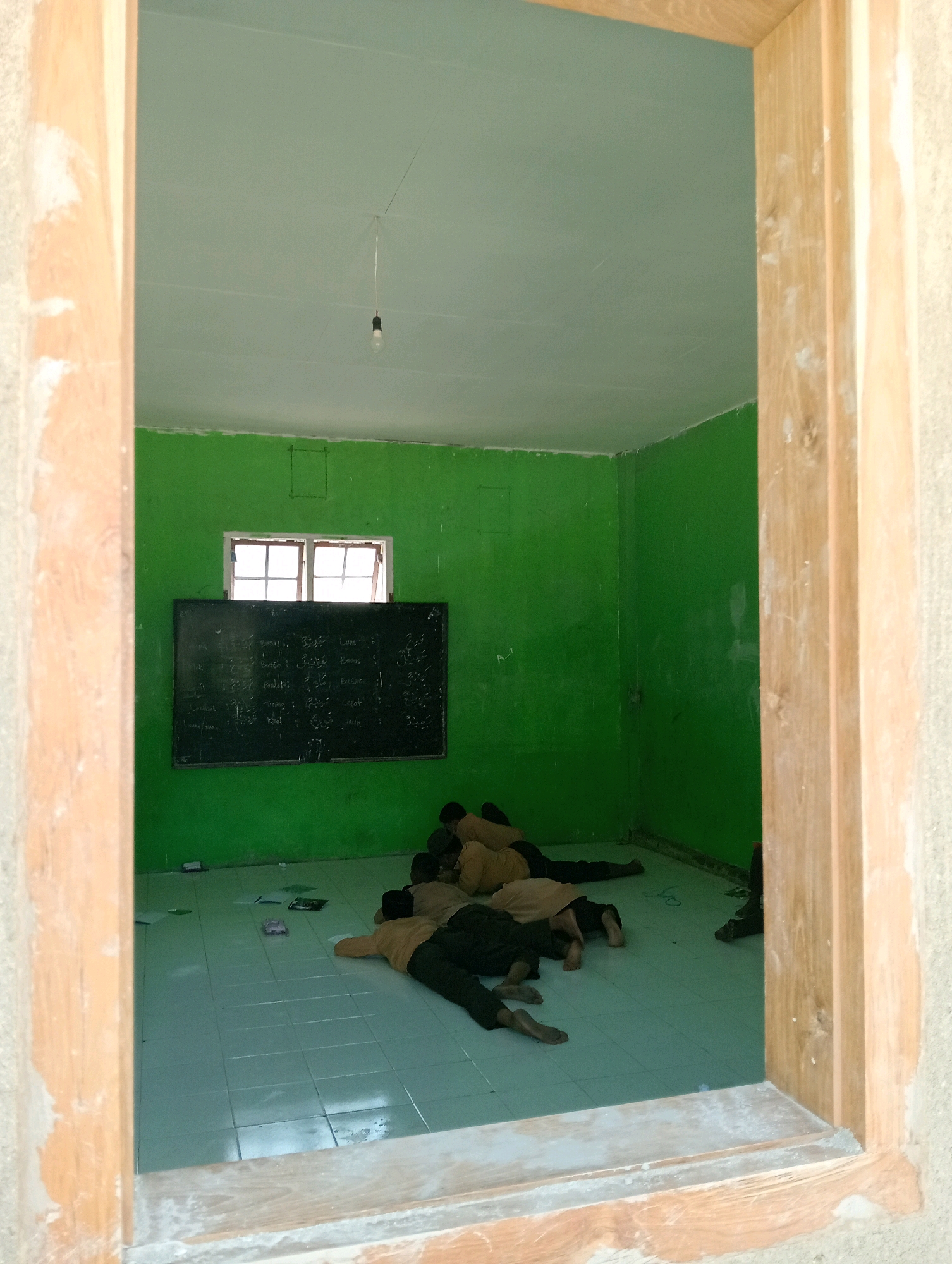 The image shows a classroom in Pagerungan Kecil, Indonesia. The walls are painted green, and the ceiling is light green. There is a window with multiple panes on one wall, above a blackboard. Several children, appearing to be students, are lying down on the tiled floor. Some are sleeping, while others may be resting. Papers and books are scattered on the floor. The scene is viewed from what appears to be an open doorway or window frame made of wood.