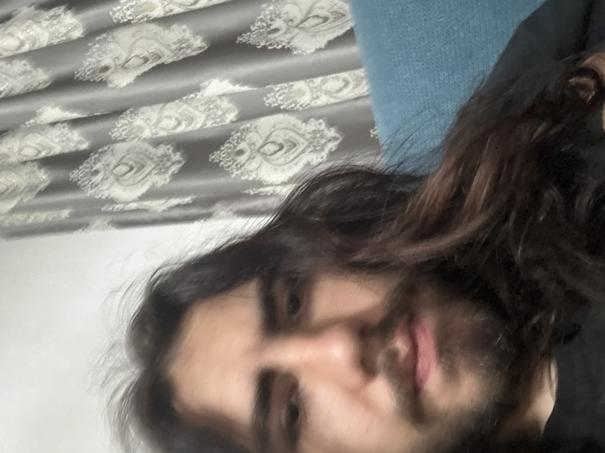 A close-up, head-and-shoulders selfie shows a young male with long, dark, wavy hair and a beard. He has dark eyes, thick eyebrows, and a light complexion. He is wearing a black shirt. In the background, to the right, is a teal-colored sofa and a light gray curtain with a white, ornate pattern. The lighting is somewhat dim. The setting is indoors.