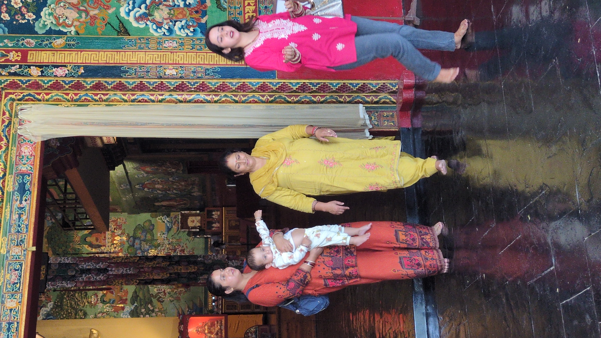Three women and a baby are in a building with colorful murals and ornamentation, possibly a temple or historic site in Fatehpur, India. From the top, a woman is lying down wearing a pink kurta and blue jeans. The next woman is wearing a yellow salwar kameez. The woman in the bottom is wearing an orange kurta and carrying a baby wearing a white pajama set. All appear to be posing for a photo, and the scene appears to be indoors.